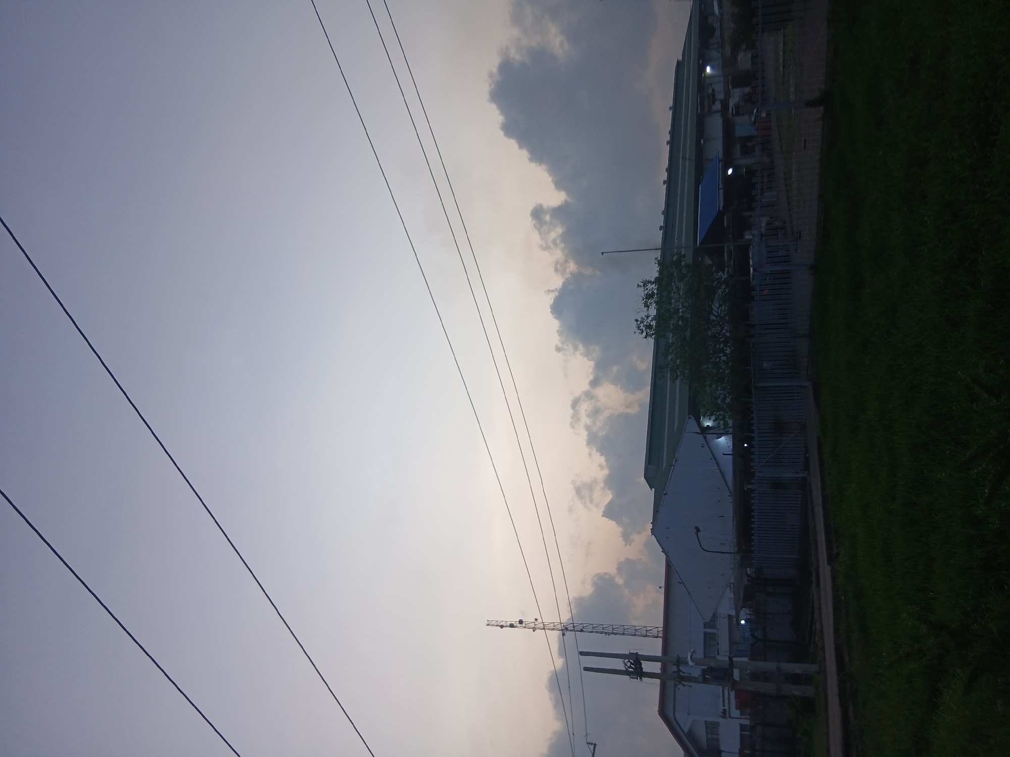 The image shows an urban scene in Lagos, Nigeria, taken at dusk. The sky is overcast with a mix of light and dark clouds. High-voltage power lines stretch across the upper left of the frame. On the right, there are buildings, including what appears to be a warehouse-style structure and smaller buildings with fences. A communication tower is visible near one of the buildings. A patch of green grass is visible along the right edge. The image is oriented sideways.