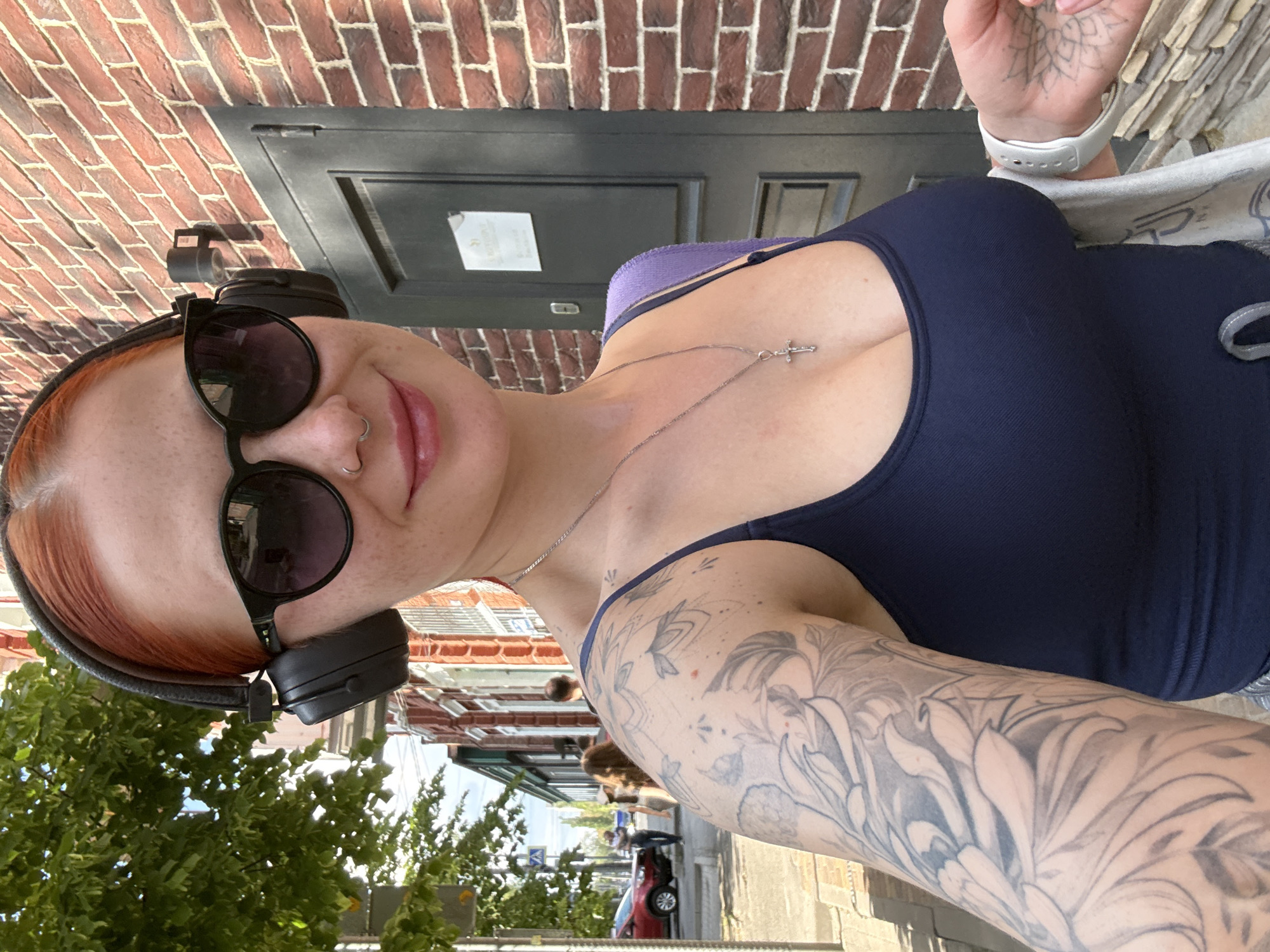 A selfie of a fair-skinned woman with red hair. She is wearing sunglasses, headphones, and a dark blue tank top. She has a cross necklace and tattoos on her arms. The background shows a brick building, a street, and trees. The location appears to be a city street, based on the presence of cars and pedestrians. Rostov-on-Don, Russia, cannot be verified from the image.