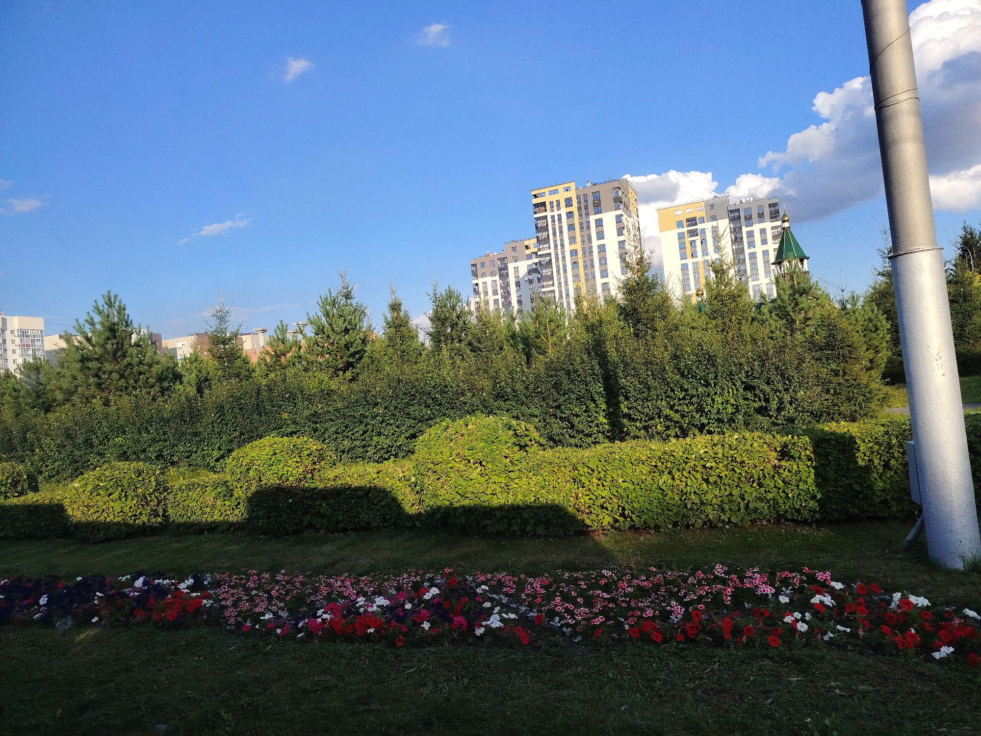 In Kemerovo, Russia, a picturesque scene unfolds with a vibrant flowerbed in the foreground, showcasing a colorful array of red, white, and pink blossoms. Beyond the flowers, meticulously manicured hedges form a lush green backdrop, casting soft shadows on the grass. Towering trees punctuate the landscape, leading the eye toward modern apartment buildings in the distance. The sky above is a brilliant blue, adorned with fluffy white clouds, suggesting a clear and pleasant day. A tall, gray pole stands to the right of the frame, its metallic surface reflecting the sunlight. The overall impression is one of urban beauty, where nature and architecture harmoniously coexist.