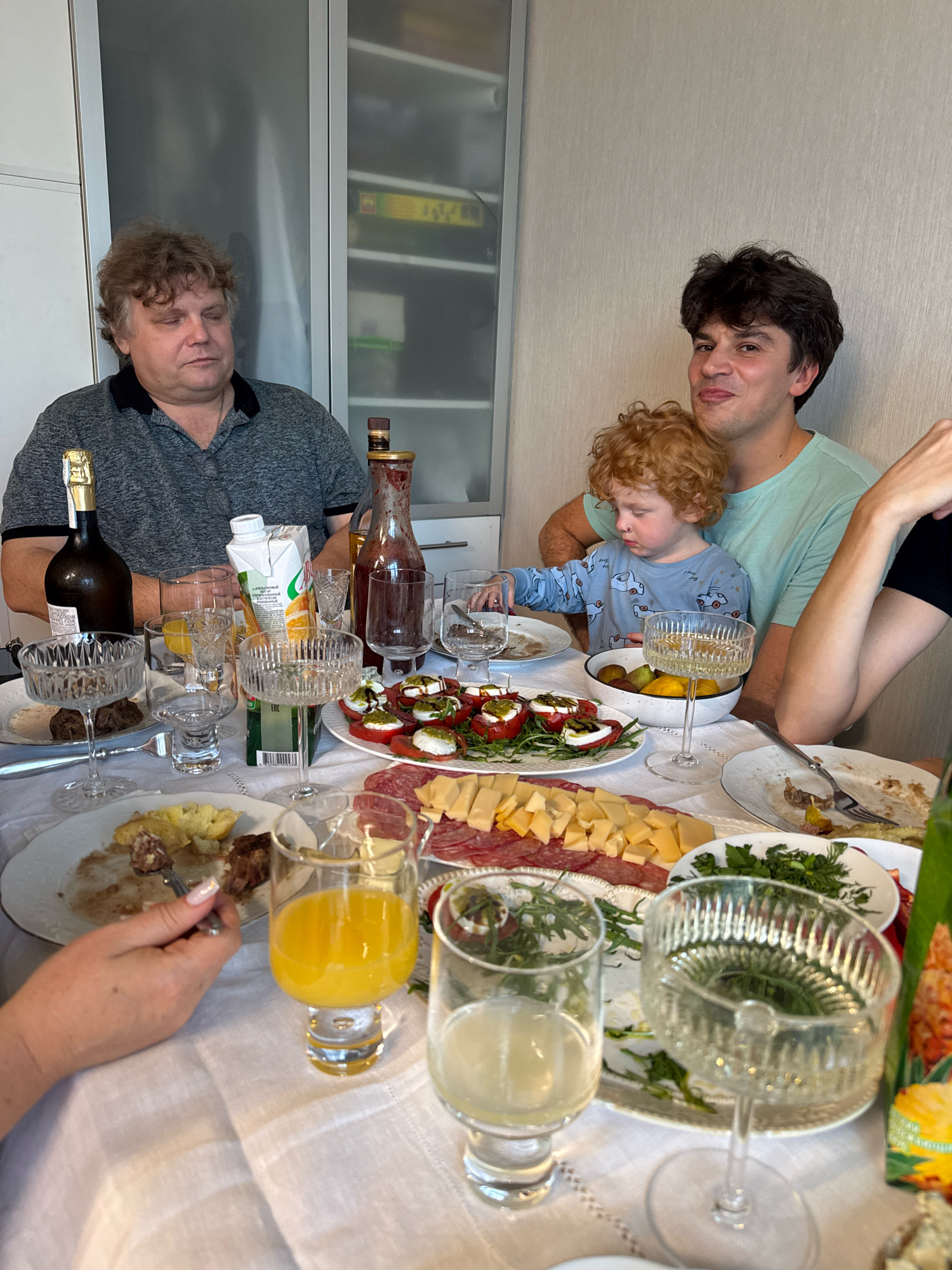 A family is gathered around a table laden with food and drinks, suggesting a meal or celebration. In the foreground, plates of food, including what appears to be sliced meat and cheese, and a salad with tomatoes and mozzarella are visible. Several glasses, some filled with yellow juice and others with clear liquid, are also on the table. A hand with manicured nails is visible holding a fork, about to eat.
On the left side of the table, a man with curly brown hair is seated, looking down. Next to him is a dark bottle of champagne or sparkling wine. Further into the frame, a younger man with dark hair is holding a young child with curly red hair. The child is wearing a light blue pajama top with car prints. The background shows a wall with some shelving and what appears to be kitchen cabinetry. The overall setting suggests an indoor meal in a home environment. The presence of a champagne bottle and the elaborate spread of food indicates a special occasion. The image is taken from a slightly elevated angle, capturing the dining table and the people seated around it. The lighting is bright and natural, illuminating the scene clearly.