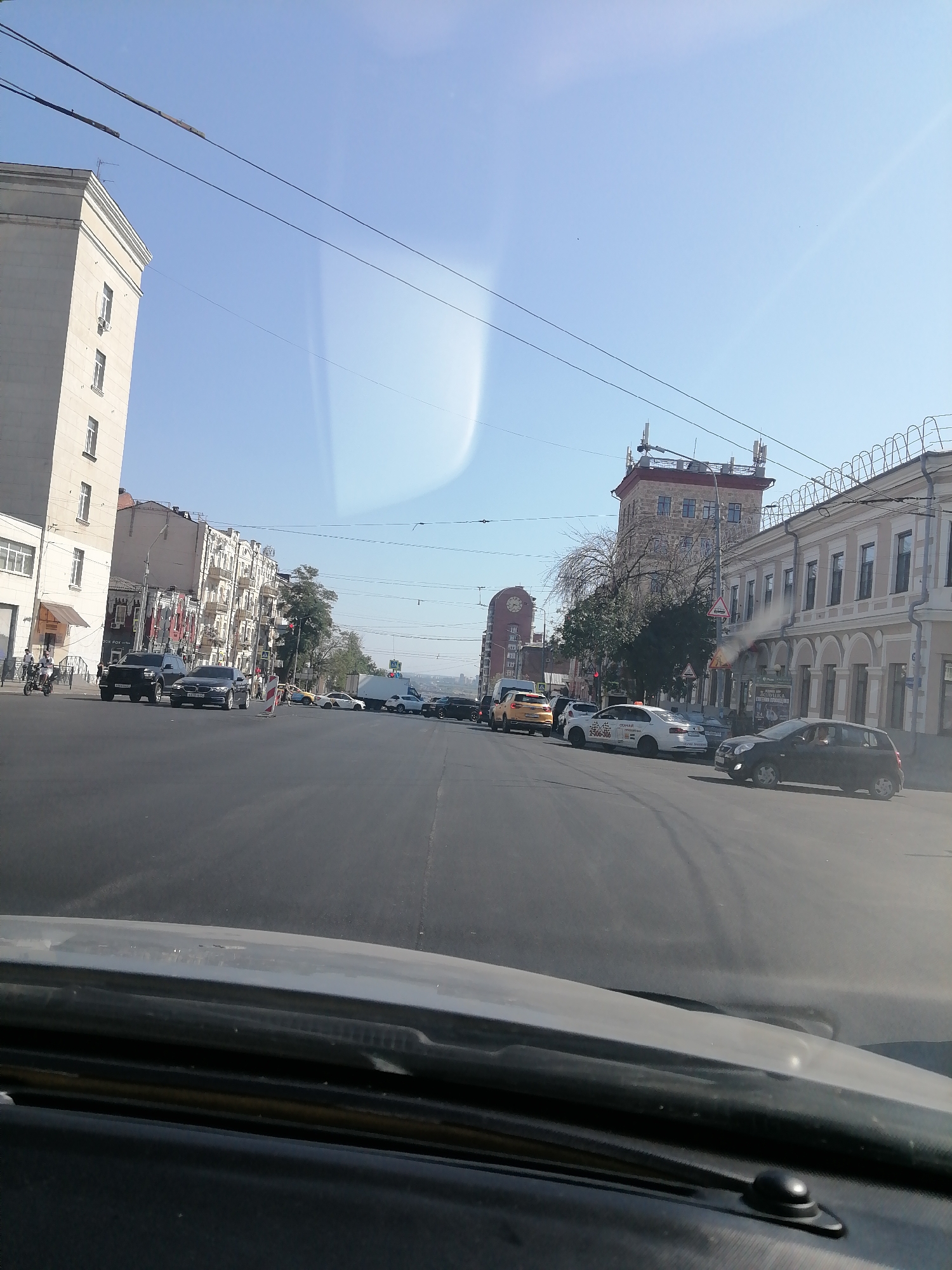 The image depicts a street scene in Rostov-on-Don, Russia, taken from the perspective of a vehicle's interior, likely the dashboard and windshield area. The road is paved and appears to be a multi-lane street. Several vehicles are visible on the road, including a black SUV, a dark sedan, a white taxi, a yellow taxi, and a white truck. Some of the vehicles are in motion, while others appear to be stopped. Buildings line both sides of the street. On the left, a tall, light-colored building with multiple windows is prominent. Across the street, on the right, a row of older buildings with decorative facades and arched windows are visible. In the background, a taller, multi-story building with a distinct clock face on one side is visible, along with a more modern-looking tower with antennae. The sky is clear and blue, suggesting a sunny day. Electrical wires and poles are present, indicating the presence of urban infrastructure. The overall impression is of a busy city street during daytime.