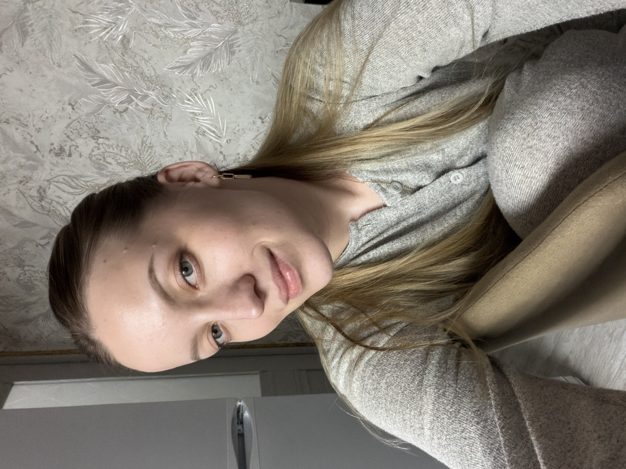 A young woman with light skin and blonde hair is looking directly at the camera. She is wearing a grey sweater with buttons. The background includes a wall with patterned wallpaper and a white cabinet. The location is not discernible from the image. No information indicates this is Reshetnikovo, Russia.