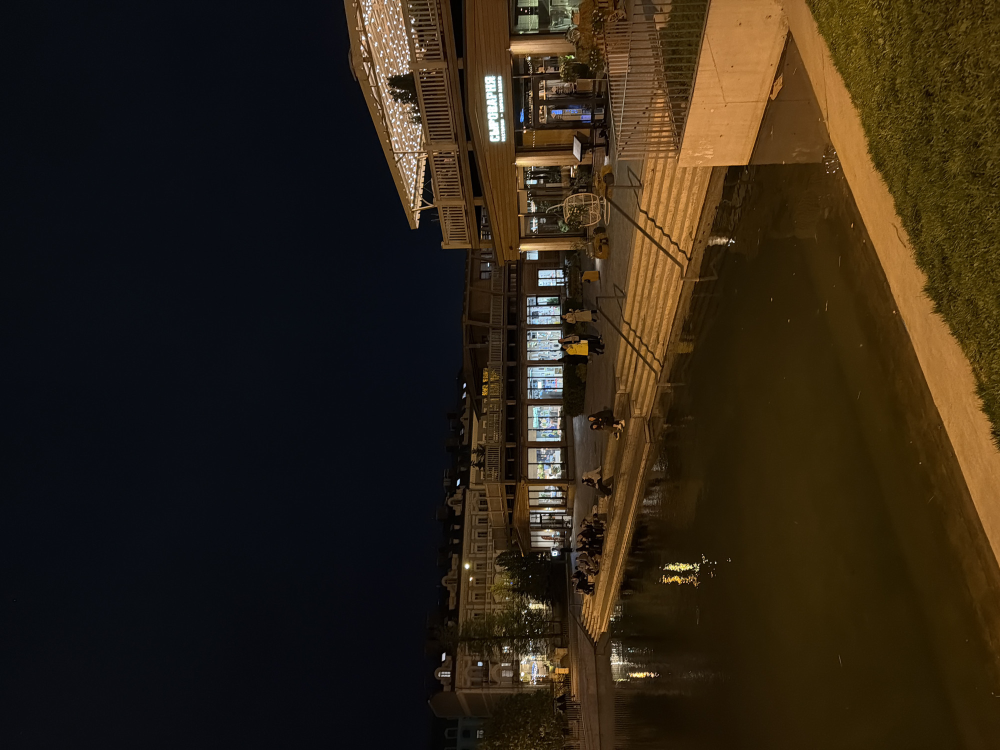 Night scene in Kazan, Russia, featuring buildings alongside a dark body of water. The buildings have illuminated windows and signs, with people visible on the steps leading down to the water. The lighting suggests a city environment at night. The right side of the image shows vegetation.