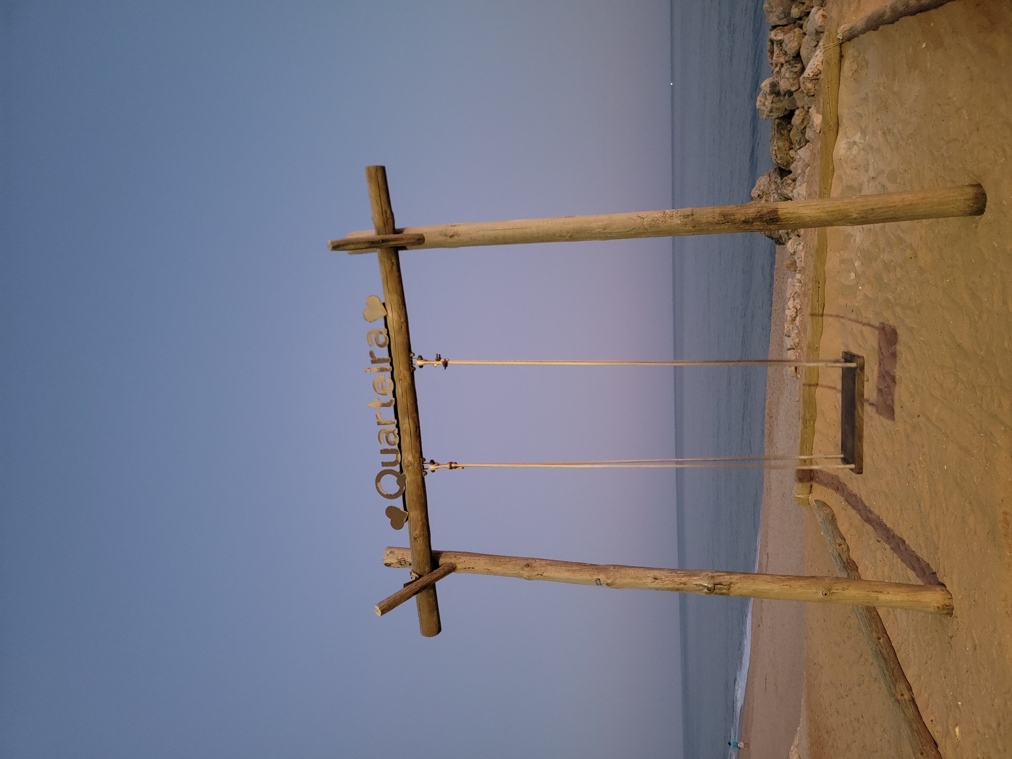 The image depicts a rustic wooden swing set on a beach at dusk. The swing set is constructed from weathered logs, with a horizontal beam supported by two vertical posts. A decorative metal sign with the word "Quarteira" and two heart shapes is attached to the left vertical post. The swing itself is suspended by ropes from the horizontal beam, with a wooden seat visible.
The setting is a sandy beach with the ocean in the background. Rocks line the edge of the beach, separating it from a paved walkway. The sky is a gradient of deep blue and lighter hues, indicating it is twilight. The lighting suggests artificial lights are illuminating the scene from the right, casting shadows on the sand.
There is no visible activity or event taking place other than the static presence of the swing and the serene beach environment. The overall mood is peaceful and tranquil, enhanced by the soft light and the quiet of the evening. The location is identified by the text on the sign as Quarteira, Portugal.