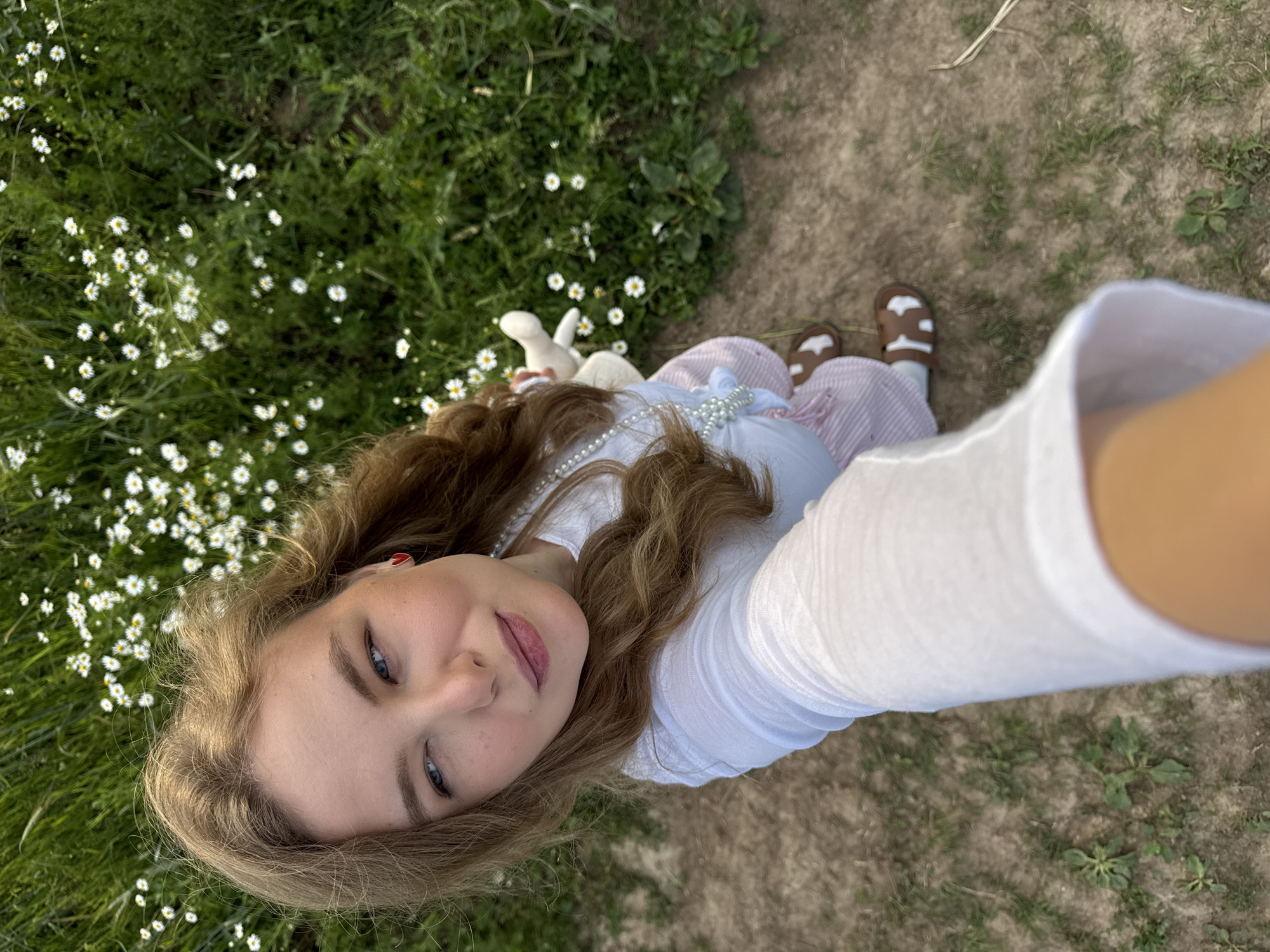 A young woman with long, wavy brown hair is lying on her back in a grassy field dotted with small white daisies. She is looking up at the camera, which is held by someone else. She has fair skin, blue eyes, and is wearing a white shirt with puffy sleeves and a pink and white striped skirt. Around her neck is a pearl necklace. She is also holding a small white stuffed animal. The background consists of more grass and some dry, bare earth, suggesting an outdoor setting, likely in a field or meadow. The lighting appears to be natural daylight, possibly late afternoon or early evening given the warm tones. The overall mood is peaceful and serene, with the woman's expression conveying a sense of calm and perhaps playfulness. The location is identified as Snovitsy, Russia.