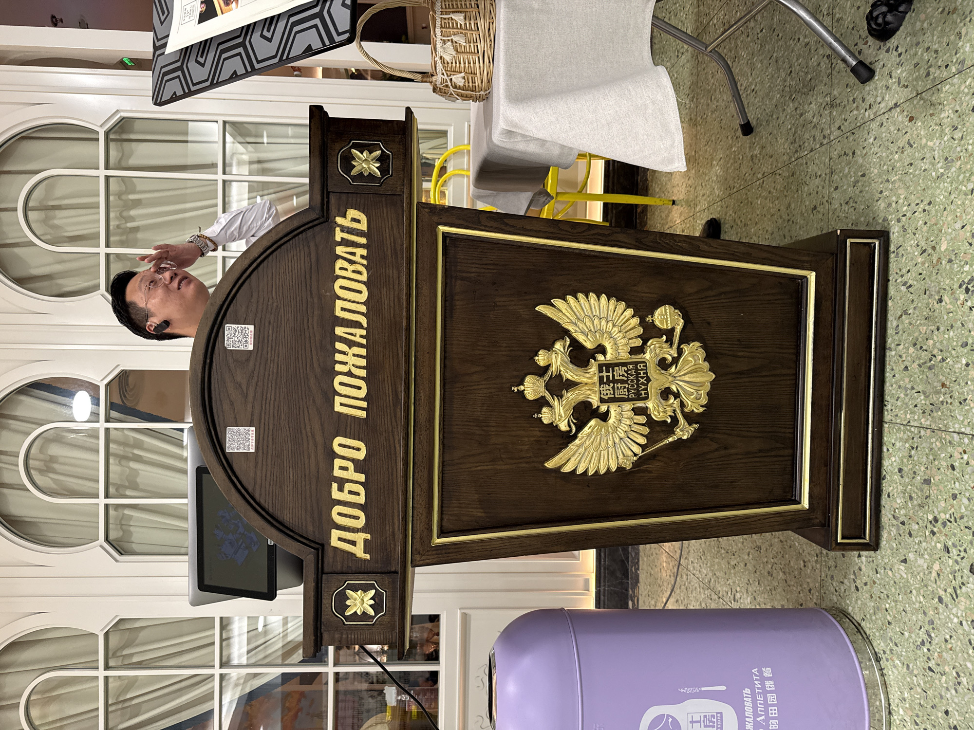 The image shows a wooden podium with a golden Russian coat of arms on the front. The podium has the words "ДОБРО ПОЖАЛОВАТЬ" in golden letters on its front panel. A man in a white shirt is standing behind the podium, partially visible, with his hand near his glasses. A screen is visible on the podium as well.
Behind the podium, a window with a grid pattern is visible. To the right of the podium, there is a purple trash bin with text in Russian, English, and Chinese, including the words "АППЕТИТА" and "РУССКАЯ НҮХНЯ", and also mentions "田园优餐".
Based on the text on the trash bin, it is highly likely that the image was taken in or near a Russian restaurant in the Tianhe District of China.