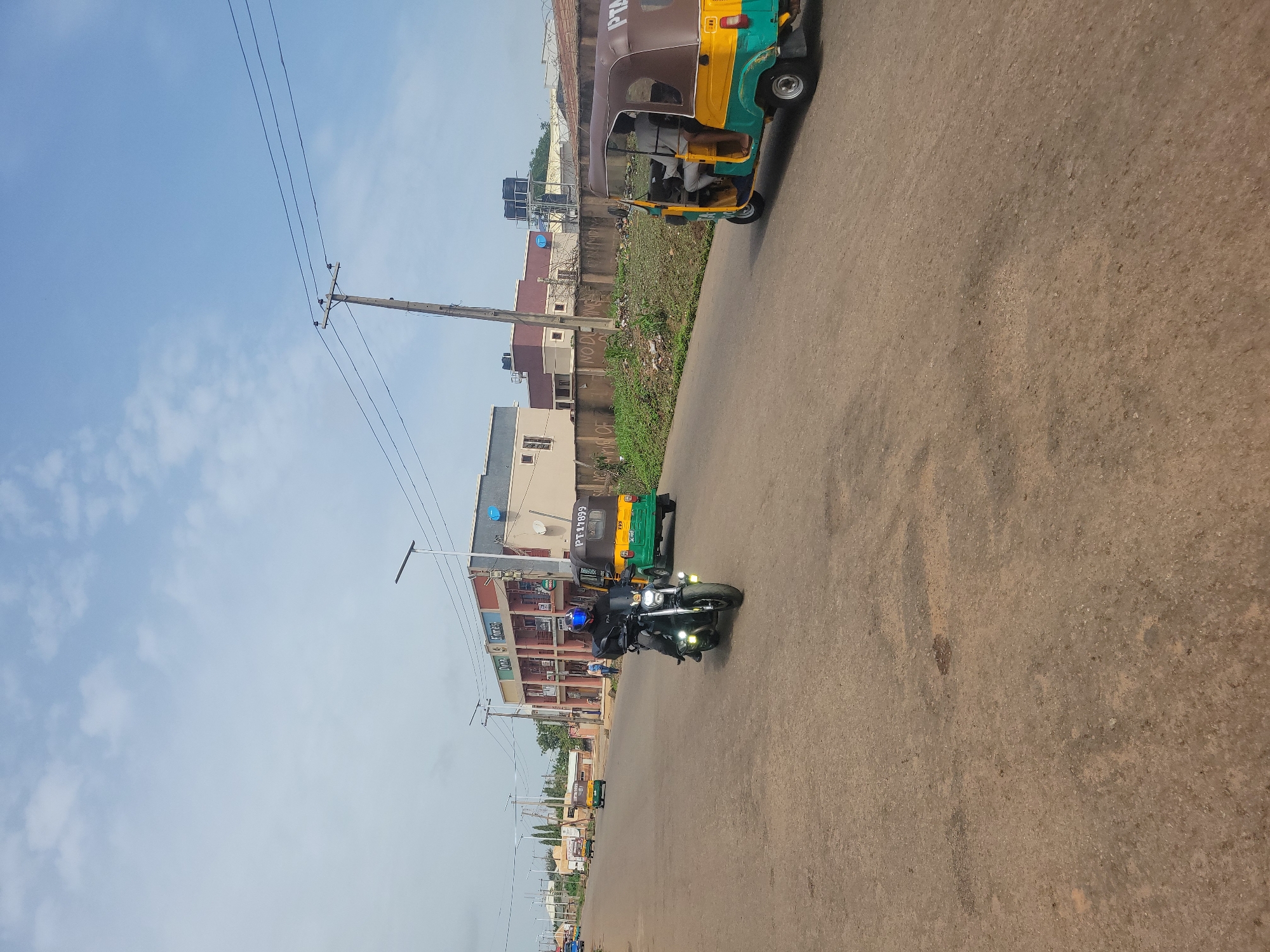 In the city of Jos, Nigeria, a motorcycle rider wearing a helmet and dark clothing navigates an asphalt road. The rider is positioned in the foreground, slightly to the left of the image's center, moving towards the viewer. Behind the motorcycle, a yellow and green auto-rickshaw, also known as a Keke Napep, is traveling in the same direction, displaying the license plate "PT-17899". Further down the road, another similar auto-rickshaw is visible. The street is lined with buildings, including a two-story structure with shops on the ground floor and apartments above. Overhead, utility poles support electrical wires, and the sky is partly cloudy with a clear blue expanse. The surrounding environment suggests an urban setting with a moderate amount of vegetation and a concrete barrier along the side of the road.