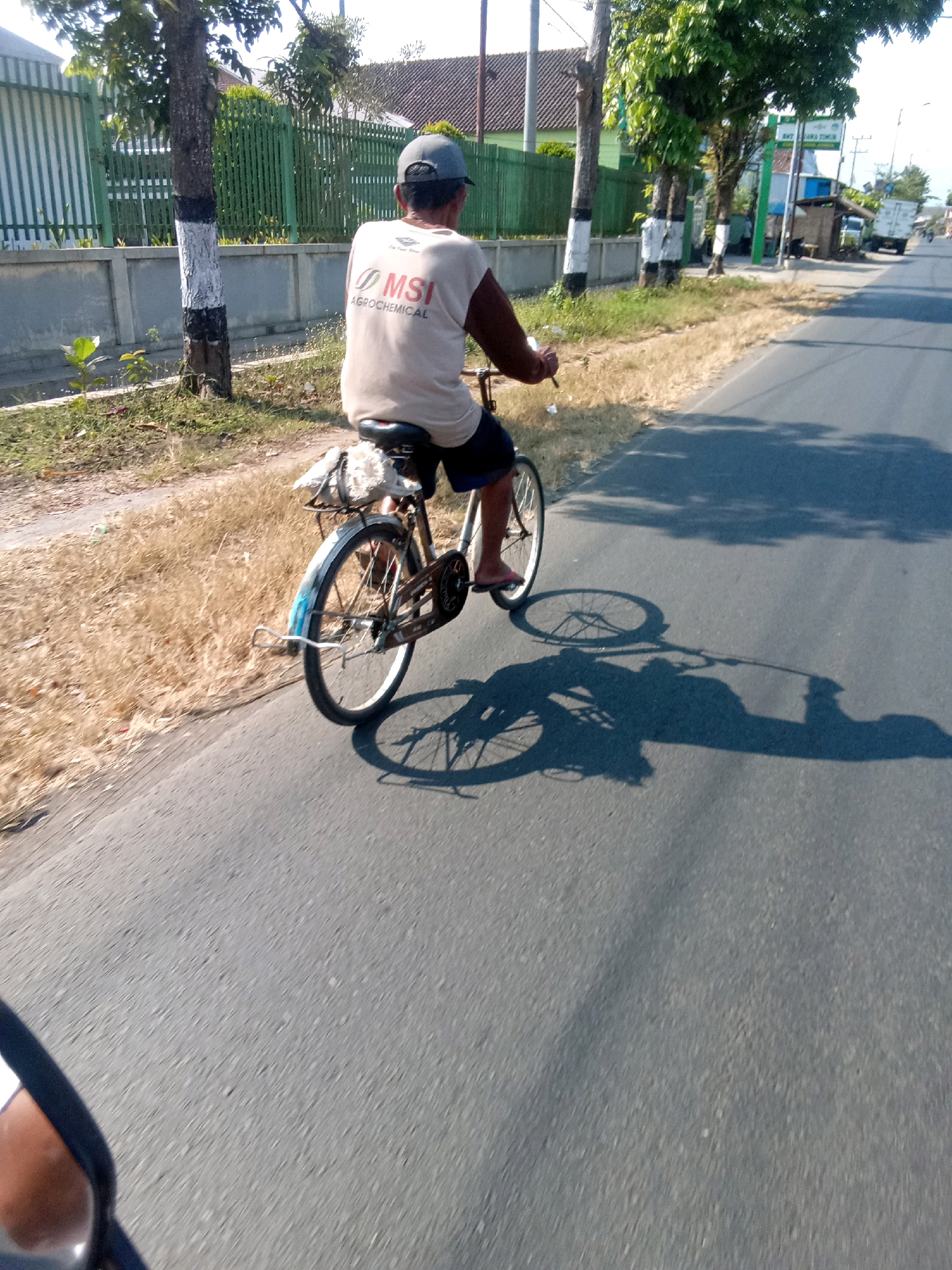 In Ajung, Indonesia, a person wearing a light-colored sleeveless shirt with "MSI AGROCHEMICAL" printed on it, and a dark baseball cap, rides a bicycle along a paved road. The rider, seen from behind and from a slightly elevated perspective, is wearing dark shorts and appears to be wearing flip-flops. The bicycle has a rear rack with some sort of white material tied to it. The road is bordered by dry, light-colored grass and vegetation on the left, and trees and some buildings in the distance on the right. The sun is casting long shadows from the rider, bicycle, and trees onto the road, indicating it is likely late afternoon. A portion of a dark motorcycle mirror is visible in the bottom left corner of the frame, suggesting the photo was taken by someone on a motorcycle.