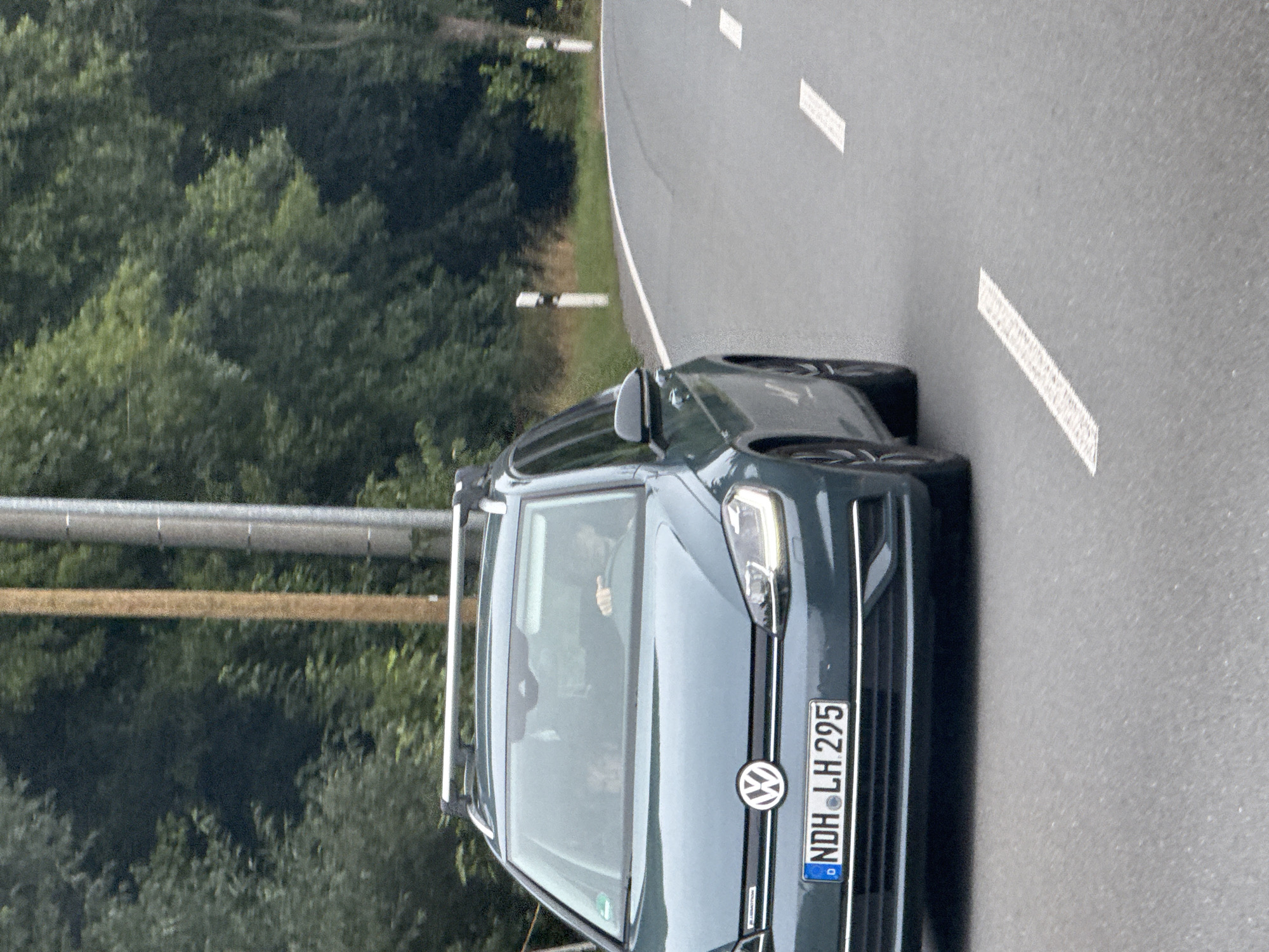 A teal Volkswagen station wagon with a roof rack drives on a paved road curving to the right. The car has a license plate that reads "NDH LH 295". Two people are visible inside the car through the windshield. The driver's hands are on the steering wheel, and a passenger is visible to the left. The road is lined with white dashes. The background consists of trees and a utility pole. The image appears to be taken in the city of Ellrich, Germany, based on the license plate format and the surrounding vegetation.