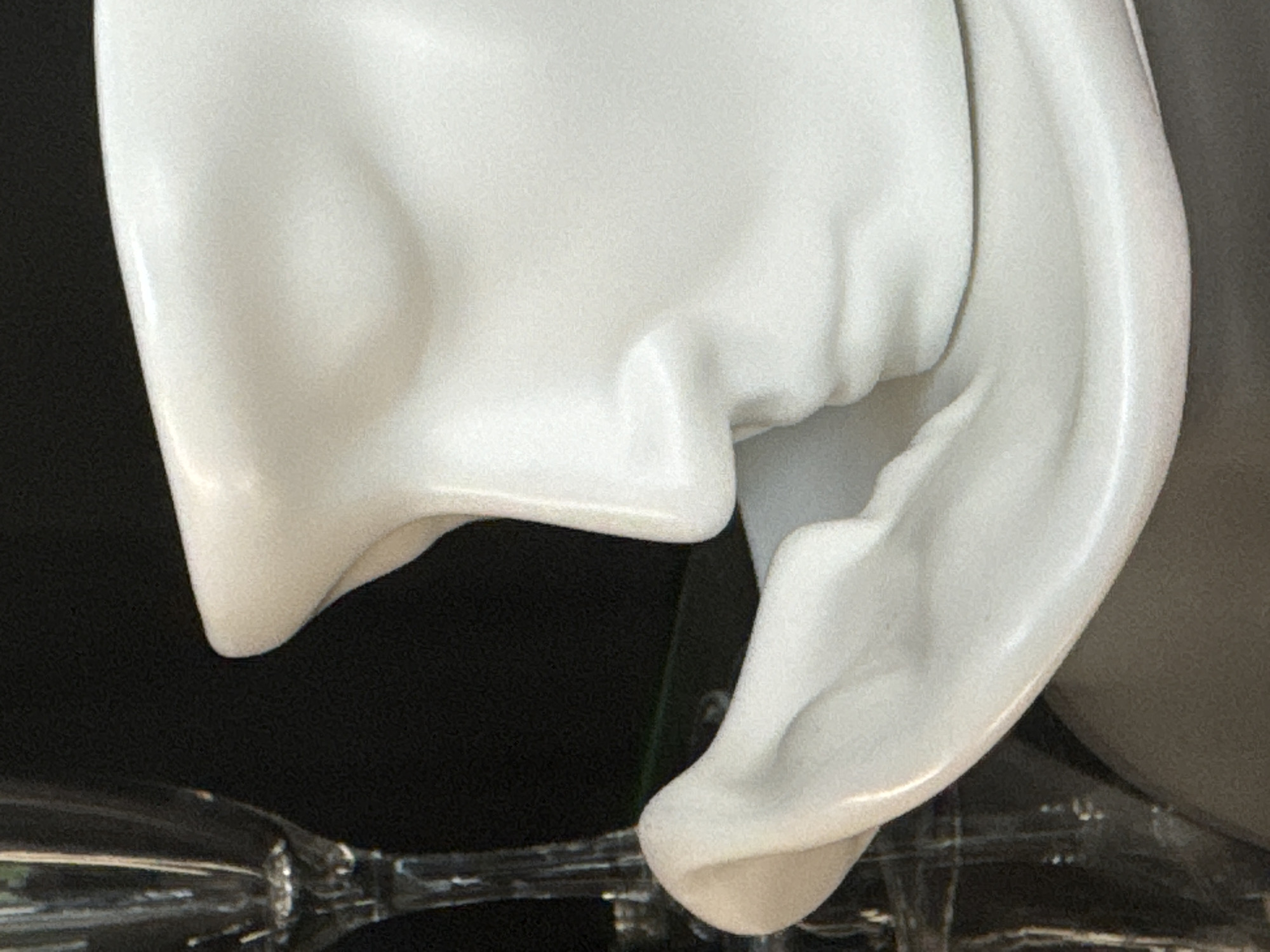 The image displays a close-up of a white ceramic object shaped like a human face in profile. The object has a prominent nose and lips, and a curved surface suggesting a cheek or chin. To the left of the ceramic face, a portion of a clear glass object, possibly a champagne flute, is visible. The background is dark and out of focus. There are no people or animals present in the image. The scene appears to be indoors, with the objects resting on a reflective surface. The lighting is soft, highlighting the smooth texture of the ceramic.
