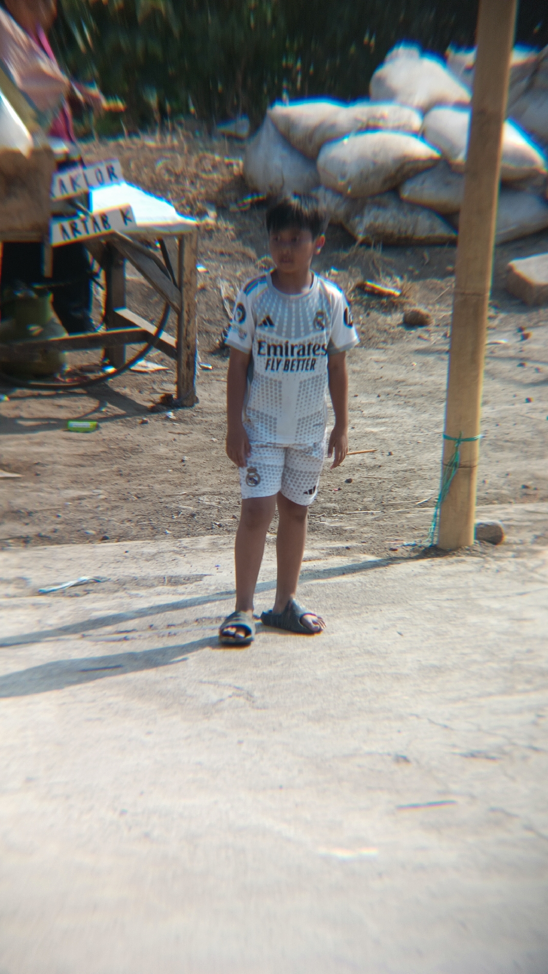 The image captures a young boy standing on a paved surface in a sunlit outdoor environment. He is wearing a white football jersey with an "Emirates Fly Better" logo and matching white shorts, and dark-colored flip-flops. In the background, there is a wooden table with some items on it, including what appear to be signs with text that is partially legible, such as "AKLOR" and "ARTAB K". Behind the table and to the right, a stack of large, white, sack-like objects are visible. A wooden pole with a green rope tied around it stands to the right of the boy. The ground is mostly dirt and pebbles, with some concrete in the foreground. The scene appears to be in Margajaya, Indonesia, given the context provided.