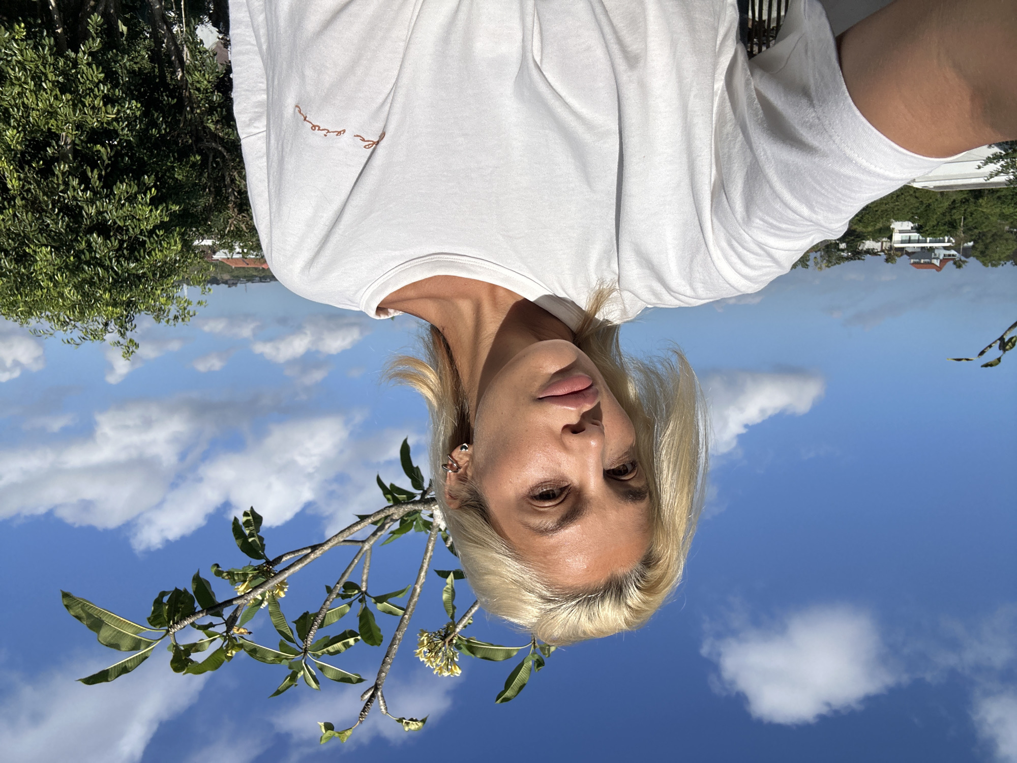 A medium shot shows a woman with blonde hair and light skin taking a selfie outdoors on a sunny day. She is wearing a white t-shirt. In the background, there is a clear blue sky with white clouds, a tree branch with green leaves and yellow flowers, and a lush green tree. Beyond the trees, several buildings are visible, including houses with red roofs. The scene appears to be set in a tropical location, possibly Kuta Selatan, Indonesia, given the vegetation and architecture. The woman's gaze is directed at the camera, and her expression is neutral. The lighting suggests it is daytime, with the sun casting shadows on her face and clothing.