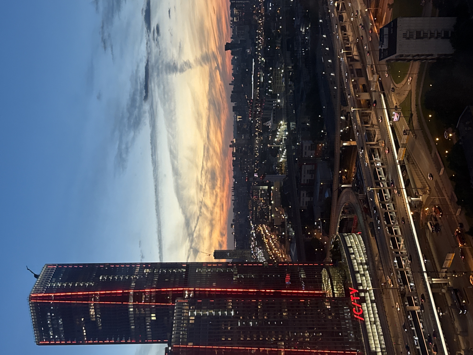 A high-angle, vertical, outdoor, long shot captures a city skyline at dusk. In the foreground, a tall, modern skyscraper with red lighting accentuates its vertical lines and illuminated windows dominates the left side of the frame. The skyscraper has a unique, angular spire at its peak. To the right of the skyscraper, the cityscape stretches to the horizon, filled with numerous buildings of varying heights. A complex network of elevated highways with flowing traffic and streetlights below the skyscraper adds depth and activity to the scene. The sky transitions from a deep blue at the top to a warm gradient of orange, yellow, and pink near the horizon, with wispy clouds creating a dramatic backdrop. The overall impression is one of urban dynamism and twilight beauty.