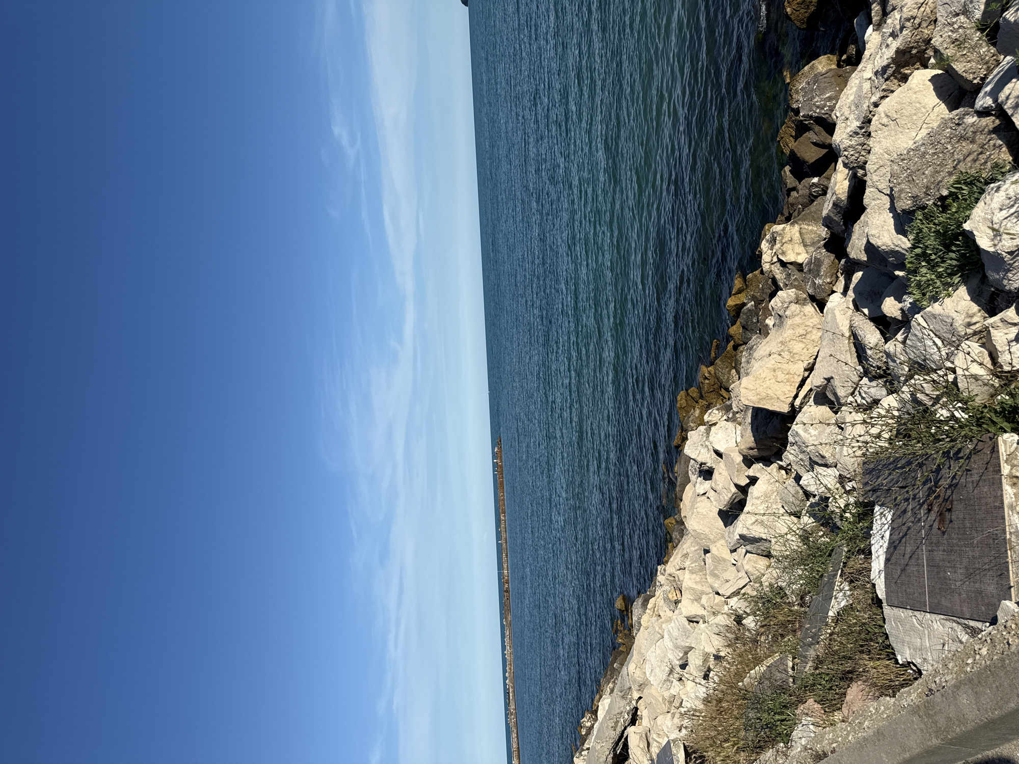 This media file depicts a seaside scene with a clear blue sky and calm, dark blue water. The foreground features a rocky shoreline composed of large, light-colored stones. Some sparse vegetation grows amongst the rocks. To the right, a section of concrete structure is visible, with a dark rectangular plaque or sign embedded in it, though no text is legible. In the distance, a long, narrow pier or breakwater extends into the sea, with small structures or posts along its length. The image appears to have been taken on a sunny day, likely in the late morning or early afternoon, given the bright lighting and clear sky. The overall impression is one of a tranquil coastal environment. The scene is consistent with a location like Varna, Bulgaria, which has a coastline on the Black Sea.