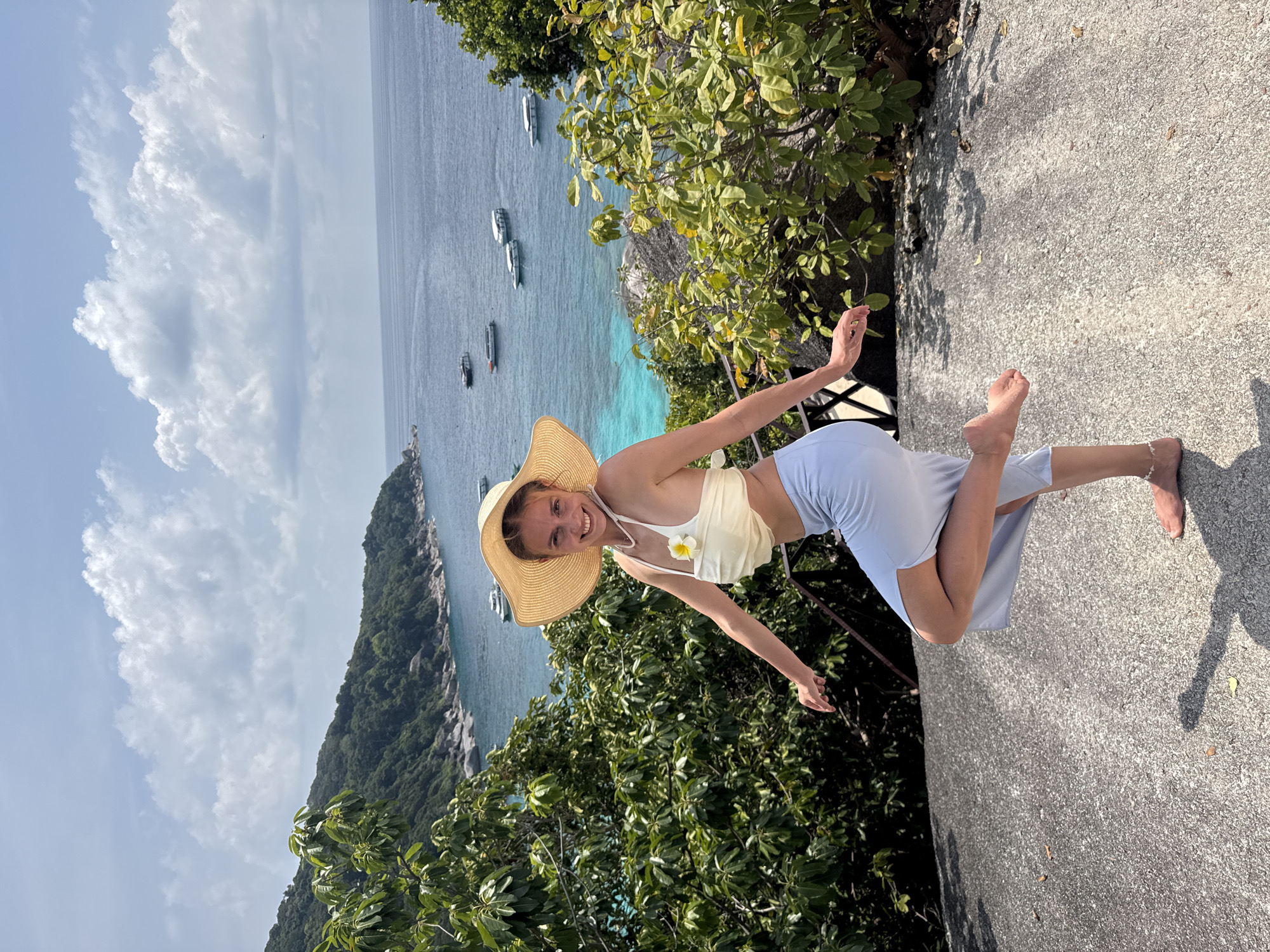 A young woman in Lam Kaen, Thailand stands on a stone surface with vegetation and the ocean as a backdrop. She wears a straw hat, a light-colored top with a flower accent, and a light-colored skirt or pants. She extends both arms outward. Boats are visible on the turquoise ocean. The sky is partly cloudy.