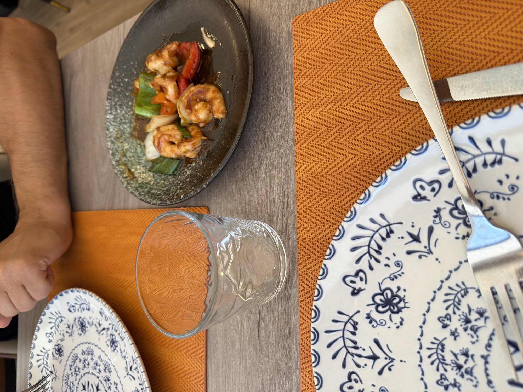 A close-up, eye-level shot shows a table setting in what appears to be a restaurant in Benidorm, Spain. In the foreground, a white plate with intricate blue floral patterns sits on an orange placemat. A fork rests on the plate, with a silver knife partially visible beneath the plate's edge. To the right of the plate is a clear, textured glass, also on an orange placemat. Further back, a dark plate holds a serving of shrimp and colorful vegetables, including red bell peppers, green onions, and white onion. A person's arm and hand are visible in the upper left of the frame, suggesting they are about to eat. The background is blurred, but hints of a dining environment are discernible. The lighting is natural, contributing to a bright and inviting atmosphere.