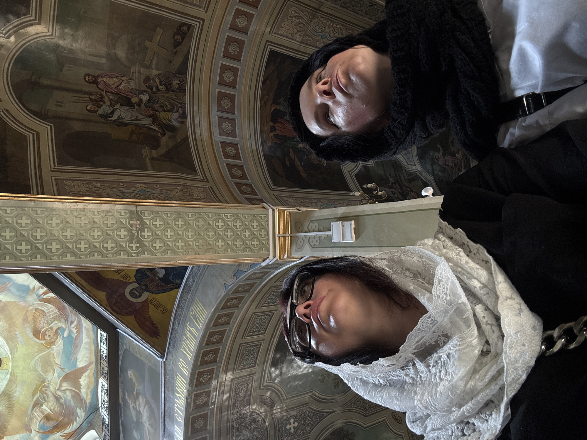 In the interior of a church, two women look upwards towards the ornate ceiling. The woman on the left wears a white lace headscarf and glasses, while the woman on the right wears a black headscarf. The ceiling is adorned with religious paintings depicting figures and scenes. The overall lighting suggests an indoor environment, possibly with natural light filtering in from high windows or a dome. The architecture of the church appears traditional, with arched structures and decorative elements.