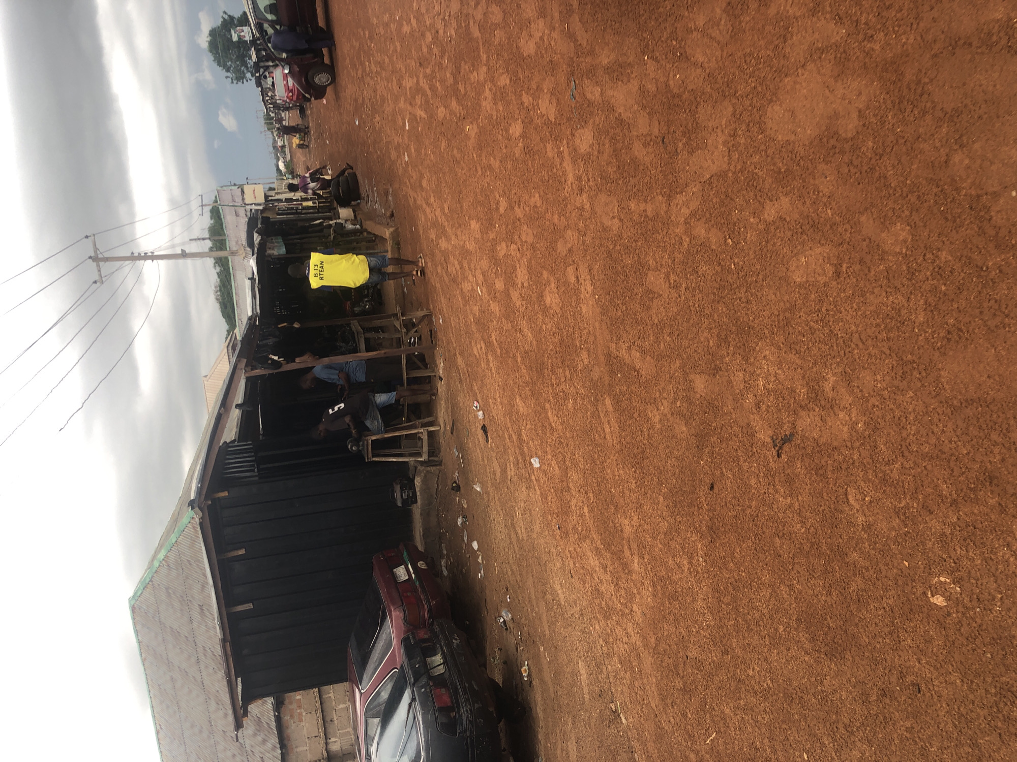 The image depicts a street scene in Wukari, Nigeria, characterized by a dusty red earth road and a row of modest structures that appear to be shops or workshops. In the foreground, the reddish-brown dirt road is uneven and scattered with debris, including plastic bags and other litter.
To the left of the image, two cars are parked. The closer car is black and appears to be older, with visible wear and tear. Behind it is a maroon sedan. Adjacent to the cars is a building with a dark, corrugated metal exterior and a weathered, green corrugated metal roof. This structure seems to be part of a commercial area.
Further down the street, there is a more active commercial area with several people present. A person wearing a bright yellow vest with "BJ3 RTEAN" printed on it stands facing away from the camera. To their left, another person sits on a stool at a wooden table, working with what appears to be a machine or tool. Another individual stands nearby, looking in the same direction. More stalls and structures line the street, with various items displayed outside.
In the background, the sky is overcast with dramatic, dark clouds, suggesting potential rain. Power lines are visible crisscrossing overhead, supported by wooden poles. A red vehicle is parked on the right side of the road, and another person in a suit appears to be interacting with it. The overall scene suggests a typical bustling, informal market or commercial zone in a Nigerian town, with evidence of both economic activity and the environmental conditions of the location.