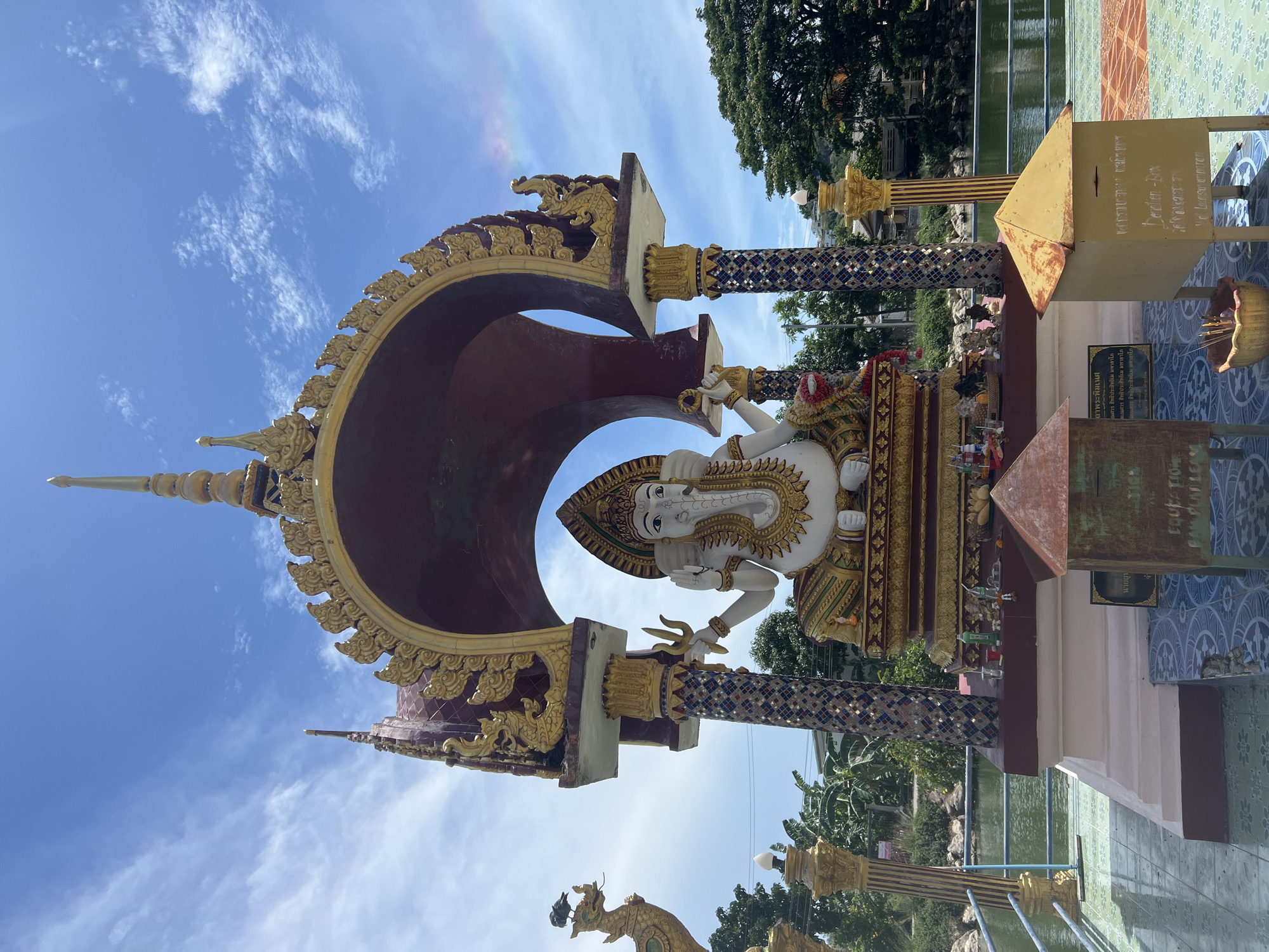 The image shows a statue of the Hindu god Ganesha in Baan Plai Laem, Thailand. The white statue is seated on a gold and red base and enclosed in a red and gold archway. The statue has multiple arms, with one holding a trident. The archway is supported by two pillars with blue and gold mosaic designs. To the right of the statue is a donation box and several signs in Thai and English. In the background, there is a body of water and lush green trees. The sky is blue with some wispy clouds. A faint rainbow is visible in the top left corner of the image.
