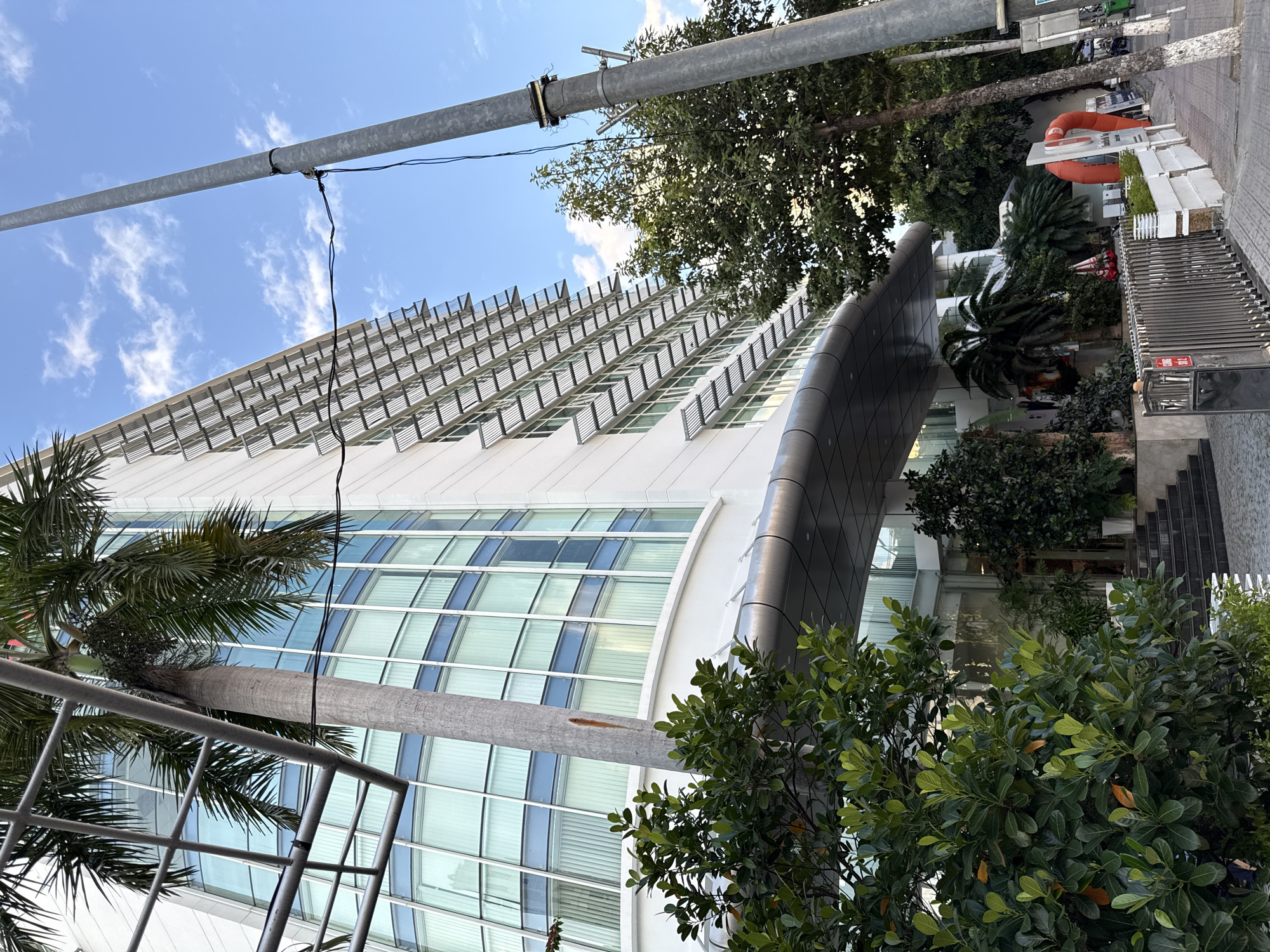 This is a low-angle outdoor shot of a modern white building with a curved glass facade, featuring horizontal louvers over each window. A palm tree is prominently positioned in the foreground, partially obscuring the building. To the right, a tall metal lamppost stands alongside a sidewalk. The building's entrance is visible, with steps leading up to a covered area. To the right of the entrance, there's a large orange life preserver and several plants. The scene is set against a blue sky with scattered white clouds. The image is likely taken in Nha Trang, Vietnam, given the presence of palm trees and the architectural style often found in Vietnamese cities. There are no people clearly visible in the image.