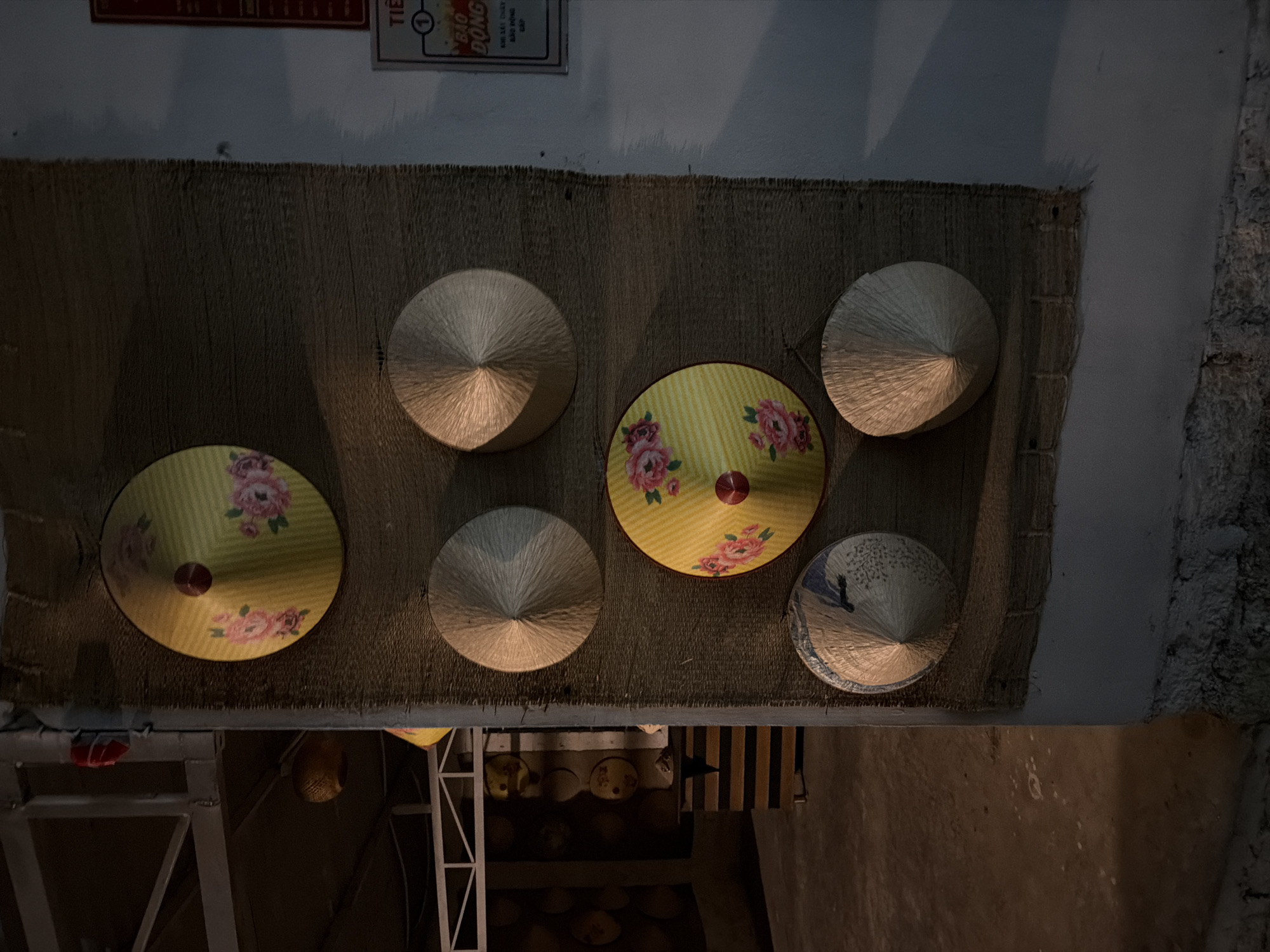 The image shows a display of traditional conical hats, known as nón lá, arranged on a textured, woven mat against a wall. Six hats are visible, with five placed prominently on the mat and one slightly lower. Three of the hats are a pale, natural straw color, showing the woven texture. One hat is decorated with a yellow background and pink floral patterns. Another hat features a blue and white design, possibly a landscape or pattern. The hats are conical in shape, tapering to a point, and have a circular brim.
The setting appears to be indoors or under a covered area, possibly a shop or market stall, given the presence of a metal shelving unit in the background with what looks like more hats or baskets on it. The lighting is dim and appears to be artificial, casting shadows and highlighting the textures of the hats and the mat. The overall mood is calm and suggestive of a cultural display.
Above the hats, a framed sign with Vietnamese text is partially visible. The text includes "TIỆN LỢI" and "BẢO ĐÔNG", and a circled number "1". The location context is Bac Nha Trang Ward, Vietnam, which aligns with the cultural significance of the nón lá. The time of day and weather are not discernible from the image.