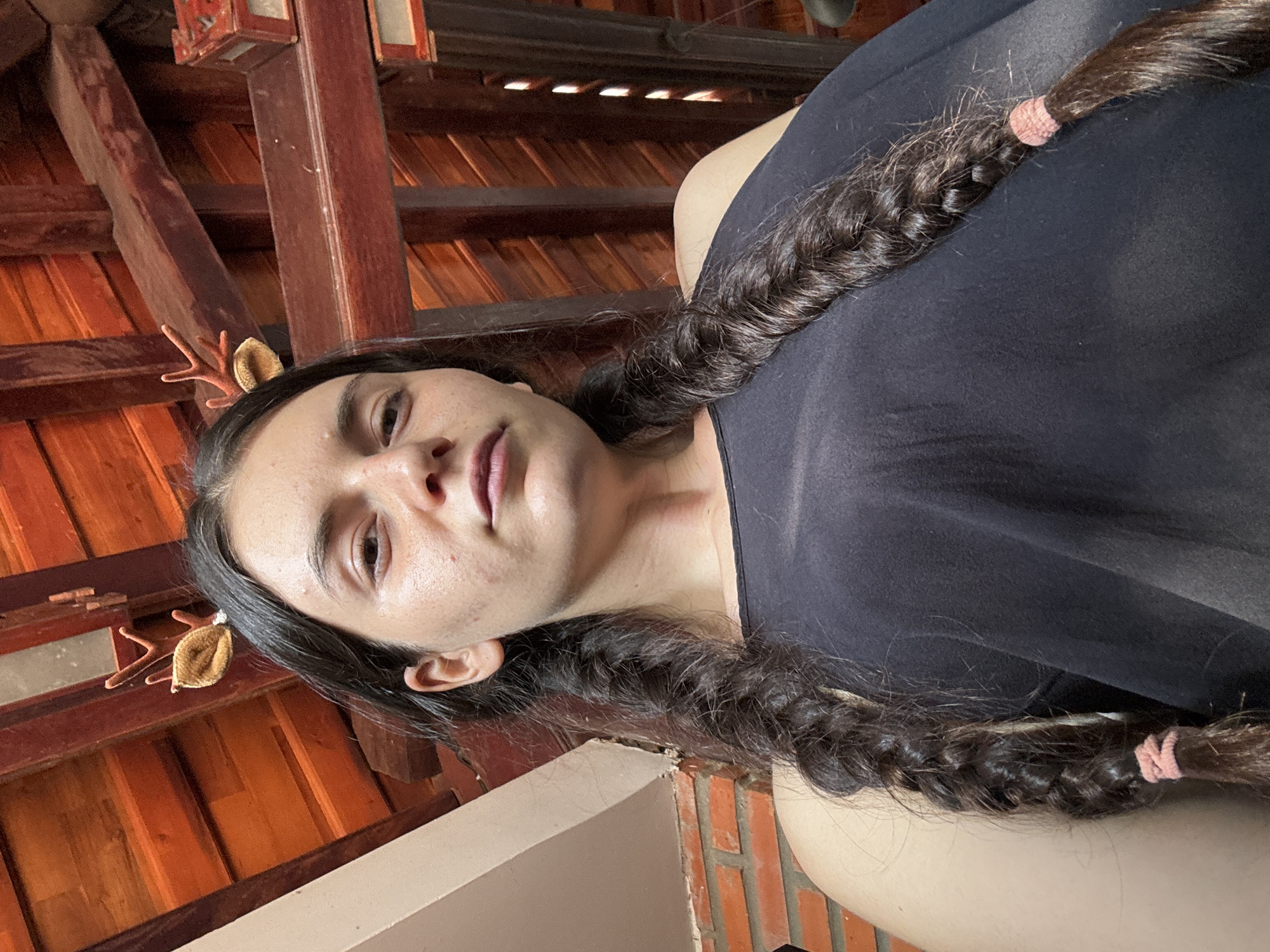 A woman with dark hair in two braids lies on her back in what appears to be an interior setting. The braids are secured with pink elastic bands. She is wearing a black top. In her hair, near her left ear, is a pair of decorative antler-like hair clips. The background consists of an ornate wooden ceiling structure, potentially indicating a traditional Vietnamese architectural style. The lower portion of the image includes a brick wall element. Given the mention of Mũi Né, Vietnam, it is plausible the image was taken in a building that reflects the region's culture.