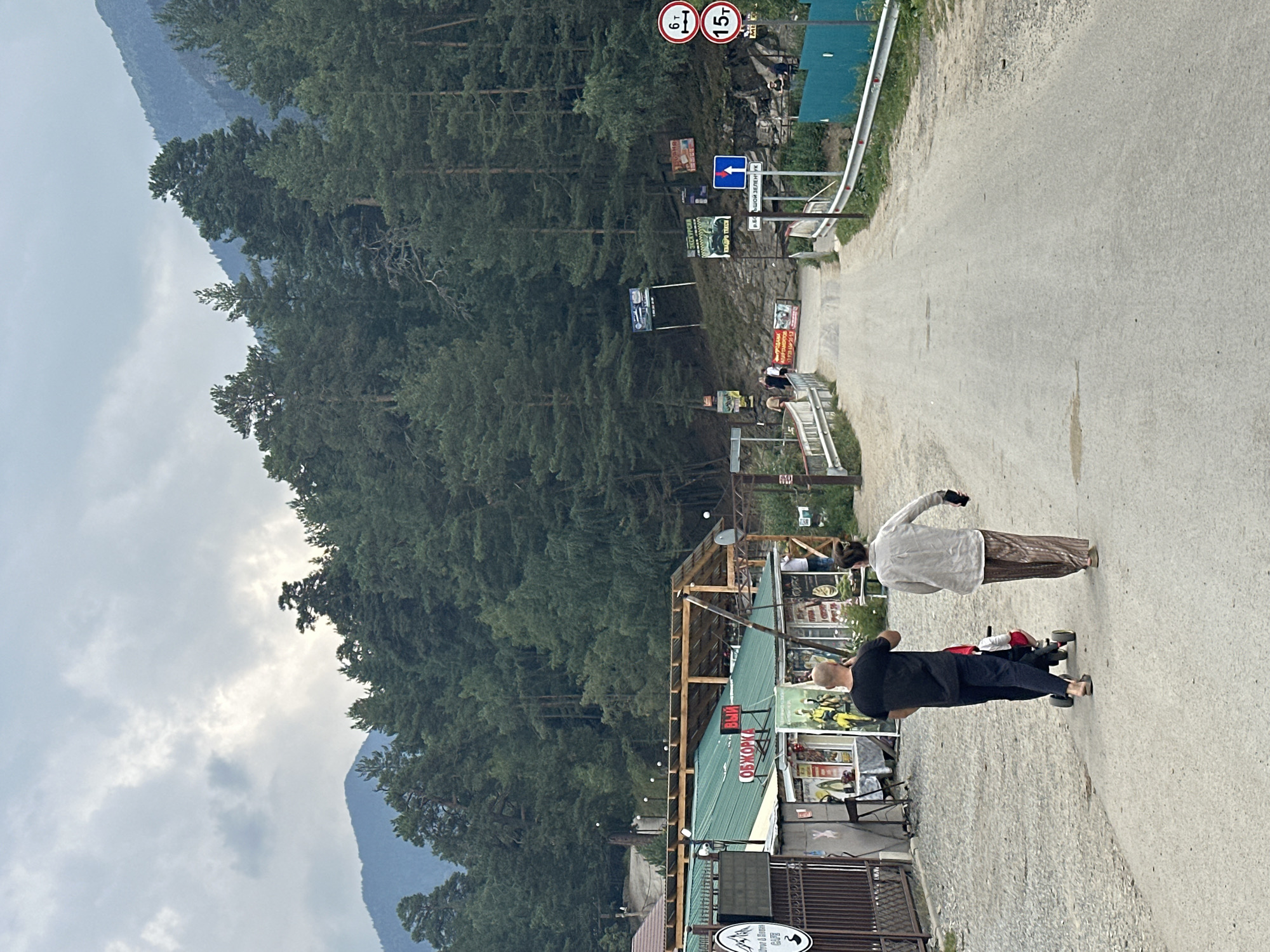 The image shows a two-lane asphalt road in the mountainous region of Arkhyz, Russia. The road is flanked by a gravel shoulder and a dense evergreen forest on the left and right. In the background, steep mountains covered in trees rise against a cloudy sky. On the left side of the road, there are several small buildings with green roofs, possibly shops or cafes, with some signage visible. One sign has a mountain and river graphic with the text "RIVER & STONE CAFE". Further down the road, there are more buildings and a bridge crossing a small stream. Several people are visible walking or standing near the buildings. On the right side of the road, there are various signs, including traffic signs indicating weight limits (6t and 15t) and a directional arrow pointing forward. Two individuals are walking away from the camera on the road, one pushing a stroller.