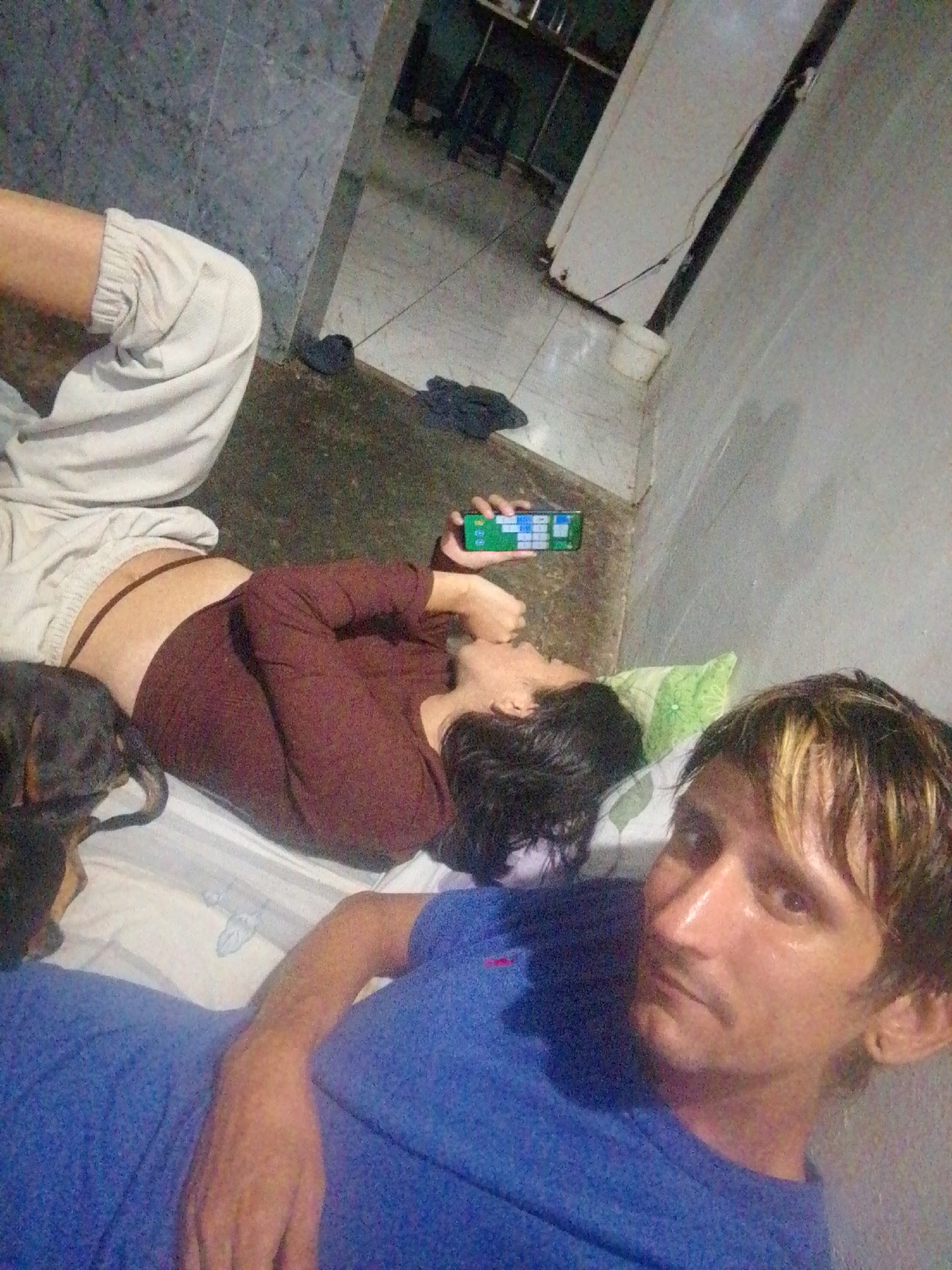 The image depicts two adults and a dog lying on a bed in what appears to be a room in a residence. One adult, wearing a blue t-shirt, is in the foreground, facing the camera. The second adult, wearing a brown shirt and light-colored pants, is lying next to them, holding a smartphone with a calculator app open. A black dachshund is partially visible on the bed, near the second adult. The room has tiled floors and plain walls. Through an open doorway, a portion of another room with a refrigerator and some furniture is visible. There is no explicit indication within the image of the location in San Carlos, Venezuela.