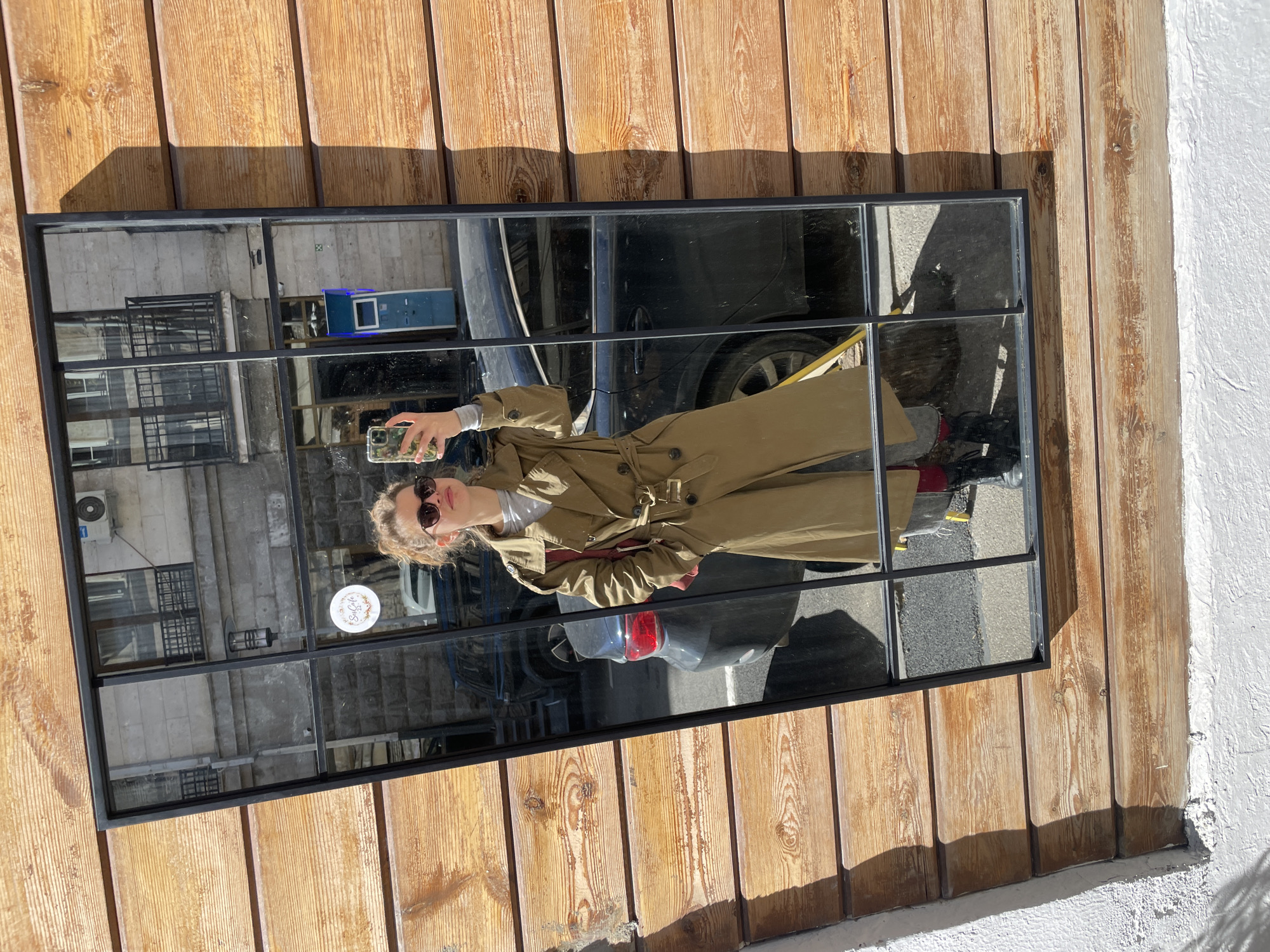 A person in a trench coat is taking a selfie in a mirror with a black frame. The mirror is mounted on a wooden wall and reflects a street scene. The person is wearing sunglasses. A car and a building can be seen in the reflection. The scene is located in Tbilisi, Georgia.