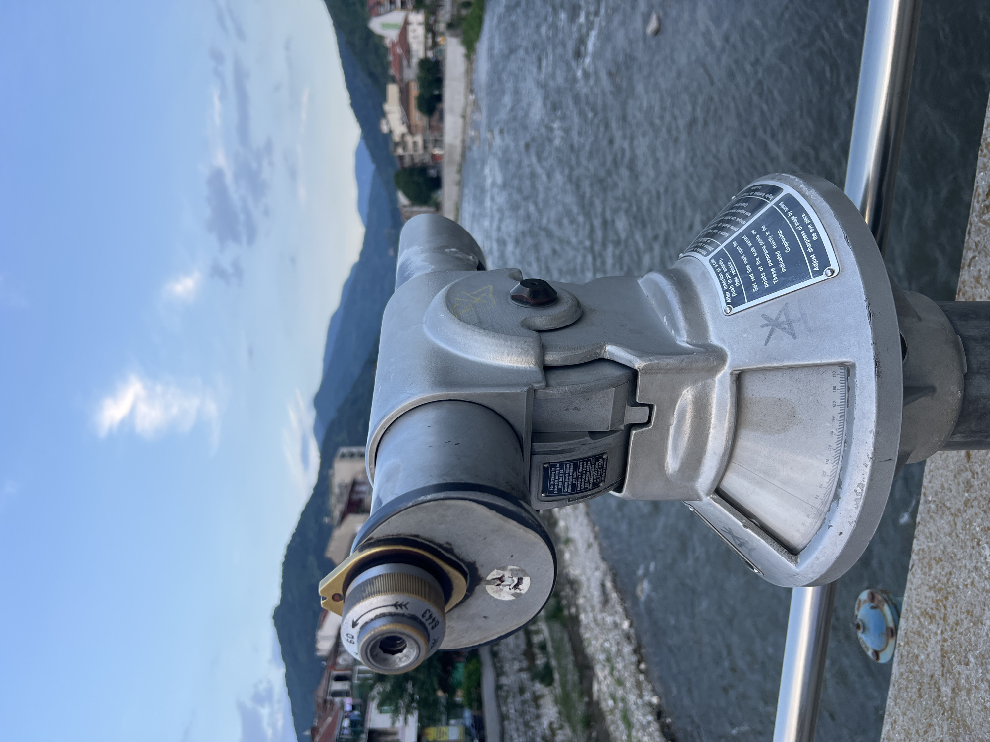 A metallic gray, coin-operated binocular viewer, likely a Graphiskop model, stands in the foreground, centered on a concrete parapet overlooking a river in Asenovgrad, Bulgaria. The viewer features an eyepiece with a golden collar and a textured focus ring marked with "60" and an arrow, along with the number "6443." A partially torn sticker is present on the side of the eyepiece mechanism. Instructional plates on the viewer's base provide operating instructions in English and another language (likely Bulgarian), detailing coin insertion, setting panorama points, and adjusting sharpness. The base also displays a horizontal scale marked with degree measurements ranging from approximately 70 to 170 degrees. To the immediate left of the viewer, a silver metal railing runs along the concrete edge.
The background reveals a flowing river, dark blue-gray, with visible rocks and pebbles along its banks, suggesting shallow areas or a low water level. On the opposite bank, a collection of multi-story buildings with various roof colors (including terracotta and darker shades) line the waterfront. Beyond the urban structures, a series of mountains rise, silhouetted in dark blue-green hues against the sky. The sky is a light blue with scattered white and light gray clouds, indicating late afternoon or early evening light conditions. No people are visible in the scene.