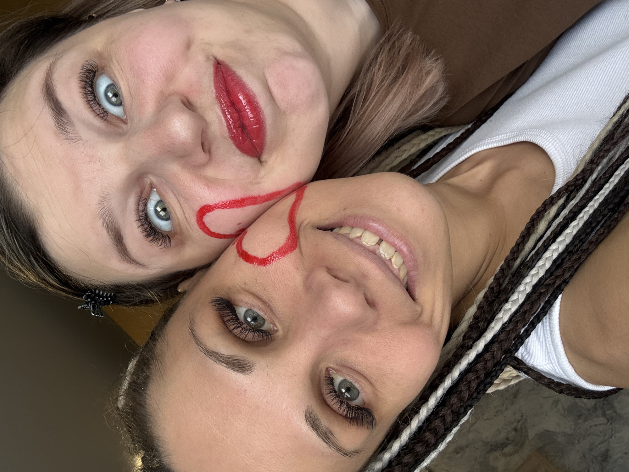 This close-up, diagonally oriented image features two women posing cheek-to-cheek in what appears to be an indoor setting in Rusanovo, Russia.
The woman on the top left has light brown hair, prominent eyelashes, and striking blue-grey eyes. She wears a brown top and has glossy red lipstick on her mouth. A bold, thick red line is drawn on her face, originating near her lips and extending onto her cheek, where it curves and seemingly connects with the other woman's face. She looks towards the camera with a relatively neutral expression. A small black claw clip is visible in her hair.
The woman on the bottom right has black, brown, and white braids and also features long eyelashes and blue-grey eyes. She wears a white ribbed tank top and is smiling broadly, showing her teeth. A similar thick red line is drawn on her cheek, appearing to complete a shape, possibly a heart or circle, formed with the line from the first woman's face.
The activity taking place is a playful photoshoot, marked by the shared red facial markings. The overall emotion conveyed is lighthearted and joyful, particularly by the smiling woman. The lighting suggests daytime indoors. There is no visible text in the image.