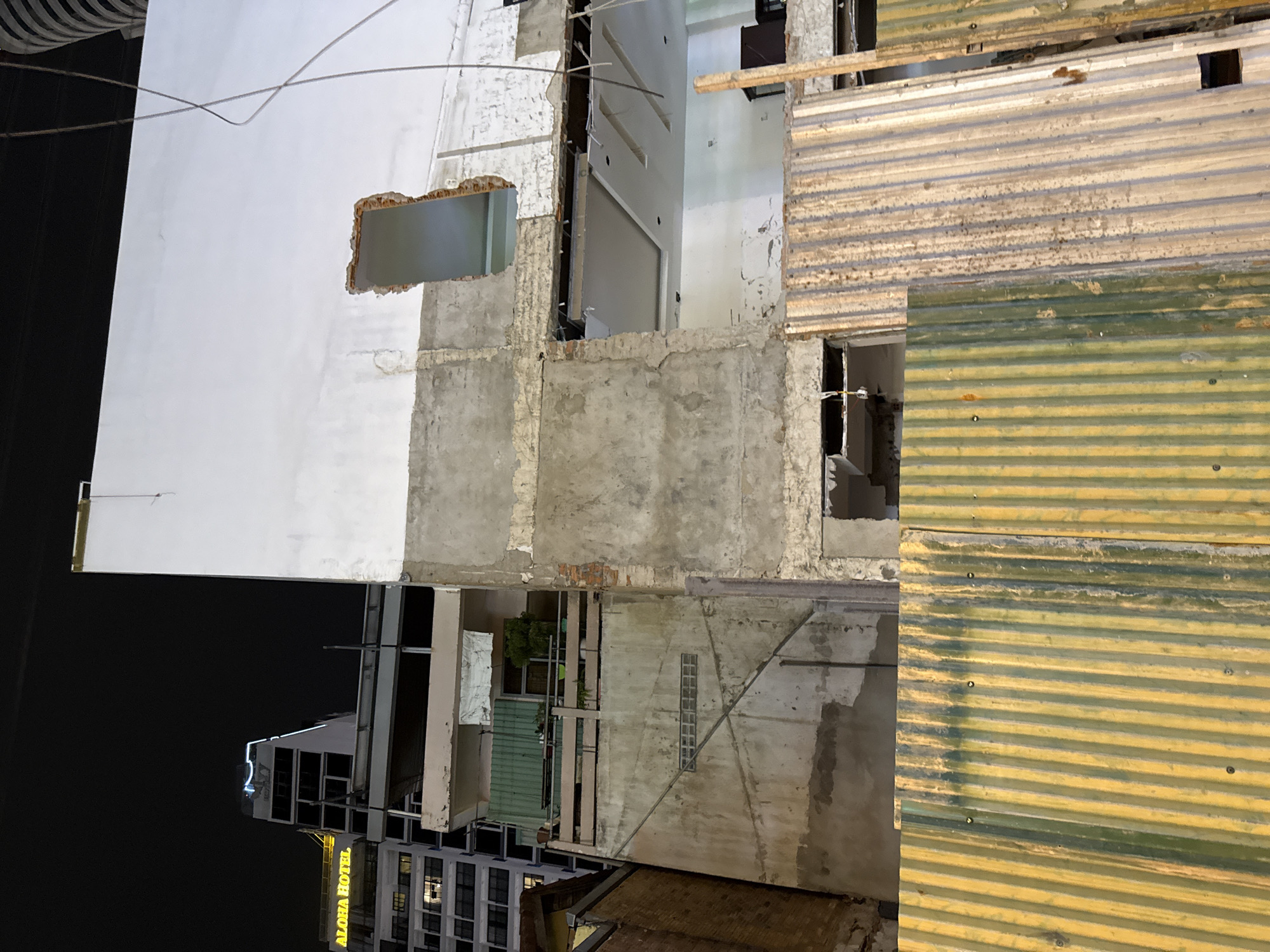This is a medium shot, eye-level, nighttime exterior photograph of the demolition of a building in Nha Trang, Vietnam. The foreground is dominated by a section of corrugated metal sheeting, colored in vertical stripes of yellow and green, some sections showing rust and discoloration. Behind this, the midground reveals the exposed, unfinished structure of the building being demolished. This includes concrete walls, exposed brickwork, and openings where windows or doors once were. One large rectangular opening on an upper level frames a glimpse of an interior space with a white dropped ceiling and lighting fixtures. Another jagged opening reveals a view into an empty space.
In the background, a modern, multi-story building with numerous windows is illuminated against the dark night sky. A sign on this building reads "ALOHA HOTEL" in yellow illuminated letters. The overall scene depicts a construction or demolition site at night, highlighting the contrast between the older structure being dismantled and the modern hotel nearby. There are no people visible in the image. The environment appears urban, characterized by the presence of buildings and construction materials.