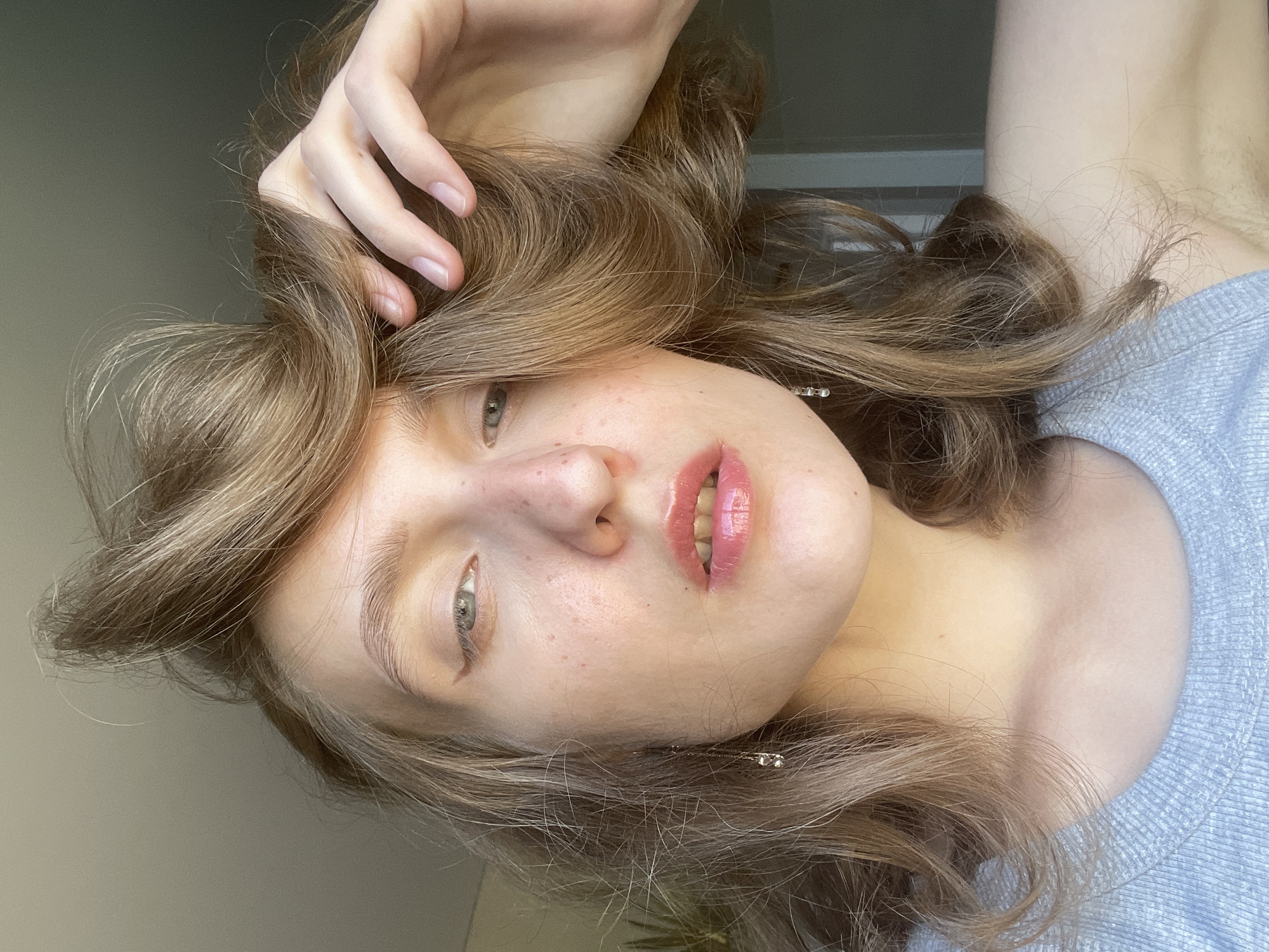 A woman with light brown hair and light skin is posing for a close-up portrait. She is tilted upside down in the frame, with her head at the bottom and her arm extending upwards. Her hand is resting on her hair. She is wearing a light blue sweater. There is a blurred light-colored wall in the background and a hint of a plant. There is no evidence to support that this image was taken in Podshivalovo, Russia.