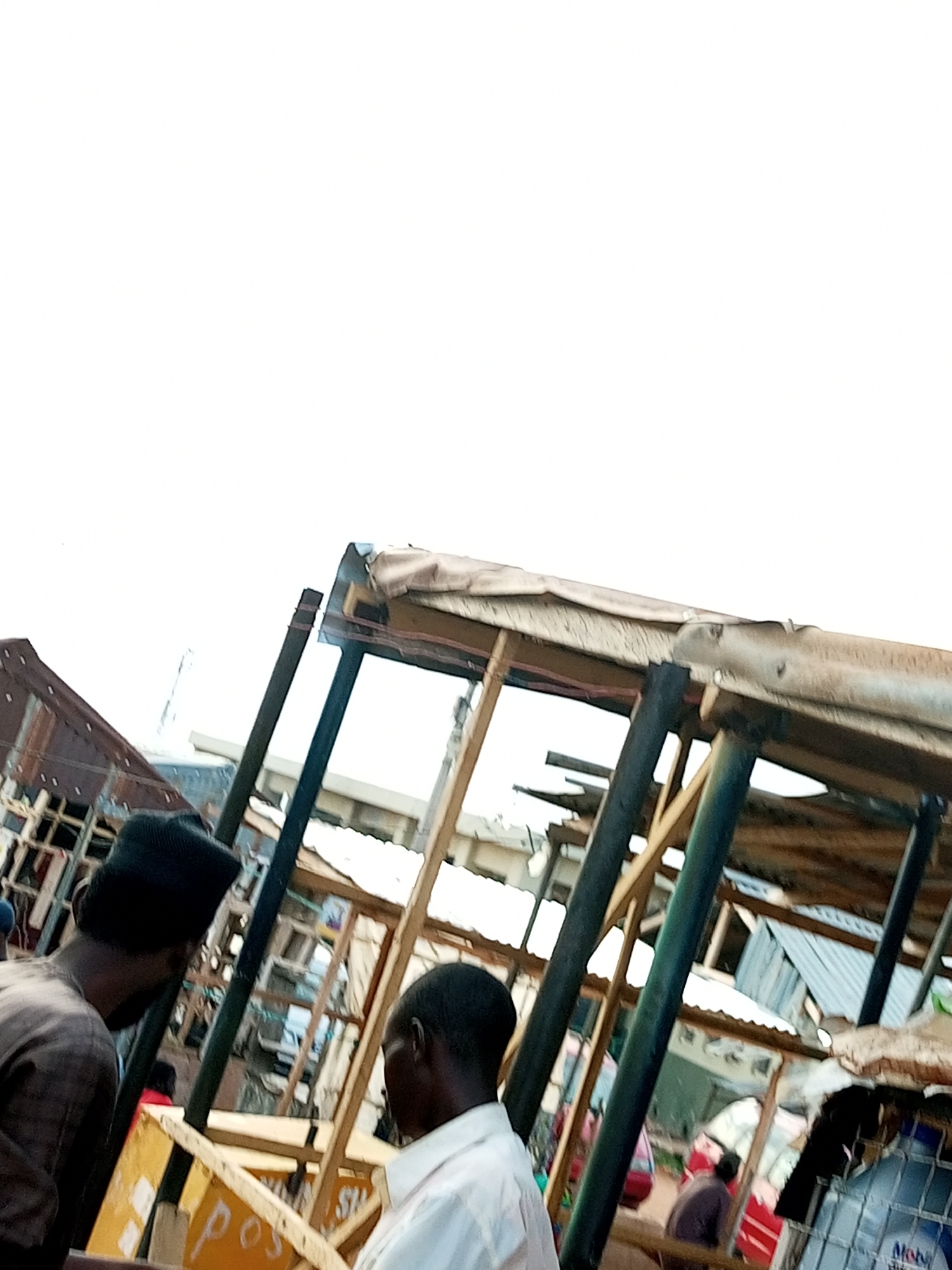 This image depicts a street scene in Gombe, Nigeria. In the foreground, two men are visible. The man on the left, wearing a dark cap and brown clothing, is seen from the side. The man on the right, facing away from the camera, is wearing a white shirt. They appear to be near a makeshift market stall with a wooden frame and a tarp covering. In the background, other market stalls constructed with corrugated metal and wood are visible, suggesting a bustling commercial area. The overall environment appears somewhat disorganized, typical of many informal markets in urban areas.