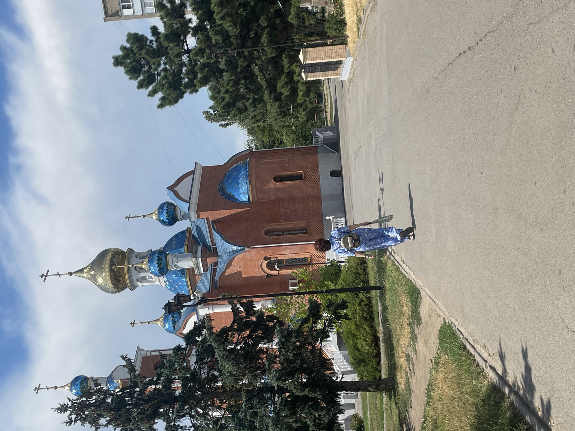 The image shows a woman walking away from the camera on a paved path, towards a red brick church with blue domes and a golden dome. The church has white trim and crosses on its domes. A tall pine tree is to the left of the church. The path is bordered by grass and some bushes. The sky is blue with some clouds. The scene appears to be outdoors in a park or courtyard area. The woman is wearing a light blue patterned jumpsuit and carrying a backpack and a bag. A tall apartment building is visible in the background to the right of the church. The location is identified as the Church of the Resurrection of Christ in the city of Azov, Russia.