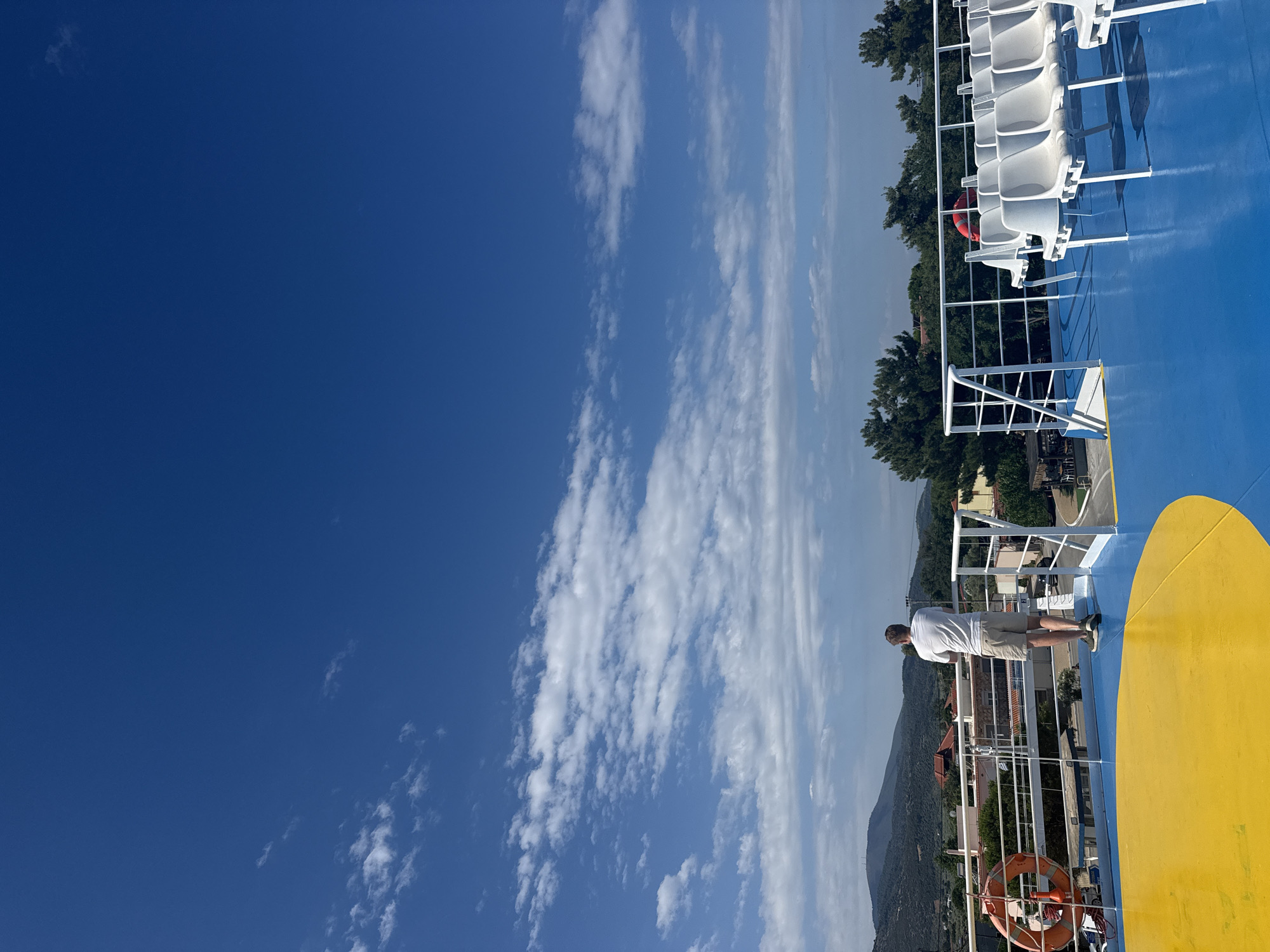 A man stands on the deck of a ferry, looking out at the landscape. He is wearing a white t-shirt and beige shorts. The deck is painted blue and yellow. In the background, there are rows of white plastic chairs, a railing, and a life preserver. Beyond the ferry, there is a view of green hills, trees, and buildings. The sky is a clear blue with scattered white clouds. The location is Prinos, Greece.