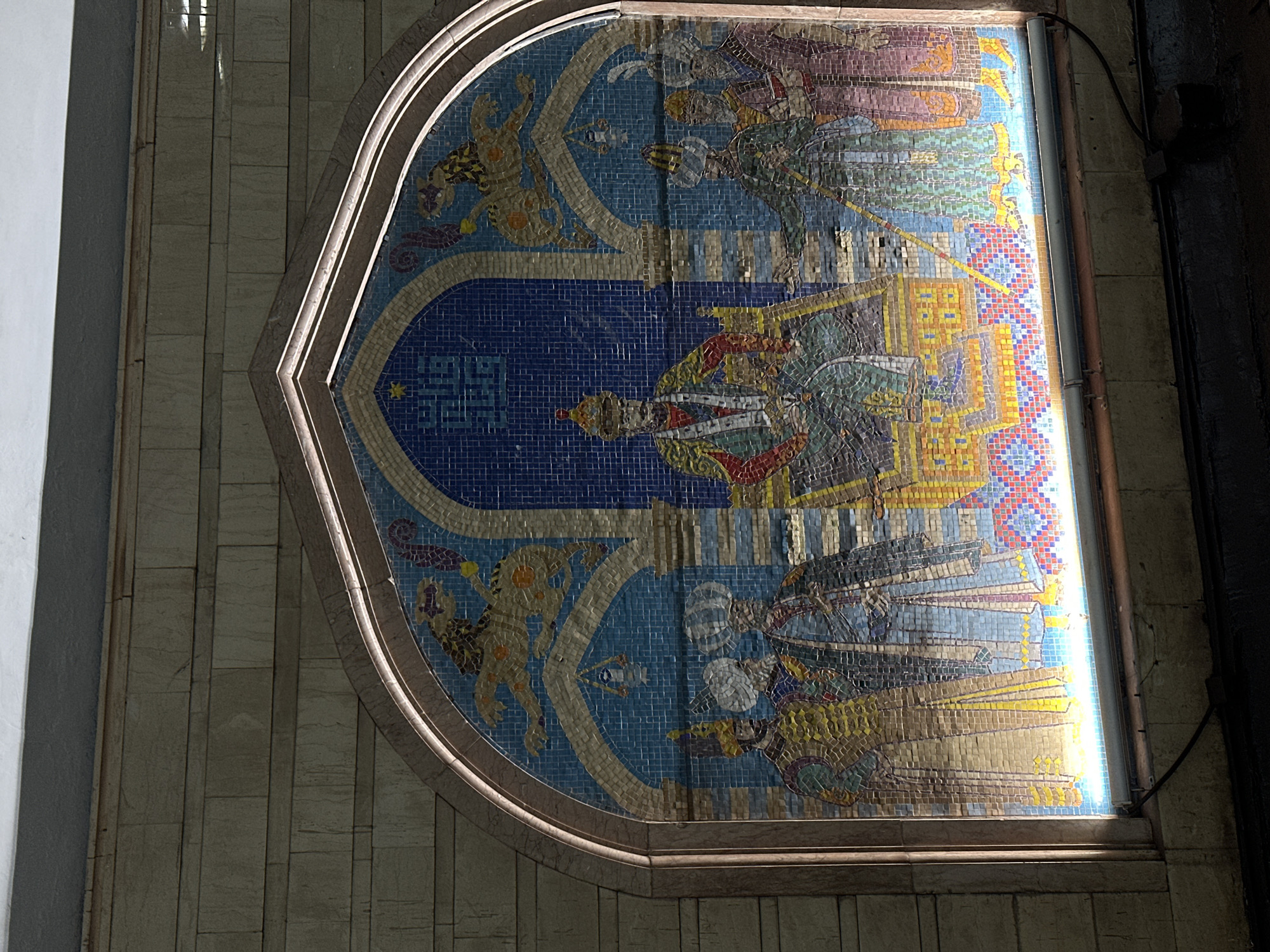 Mosaic art in an architectural setting in Kazan, Russia. The mosaic artwork is set in a non-rectangular space resembling an arch. The artwork itself depicts a king or ruler on a throne, surrounded by figures on either side. The mosaic's design includes symbolic elements like zodiac or mythical creatures. The artwork is illuminated by a linear light source. The setting appears to be an interior of a building with tiled walls.