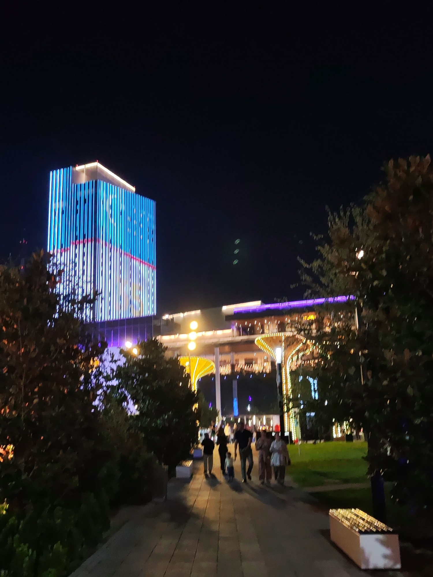 The image depicts a nighttime scene in Tashkent, Uzbekistan, likely within a modern urban development or park area. In the background, a tall skyscraper is illuminated with vibrant blue and white vertical lights, with a section near the top featuring red and white lights. Adjacent to the skyscraper, a lower, multi-story building is also lit, with purple lights along its upper edge and numerous smaller lights suggesting a commercial or entertainment venue. In the foreground, a paved walkway is visible, bordered by trees and green lawn areas. Several people are walking along the path, their figures silhouetted against the illuminated background. One person appears to be walking a small child. A bench-like structure, possibly a planter, is present along the right side of the walkway. The overall atmosphere suggests a lively evening in a contemporary city environment.