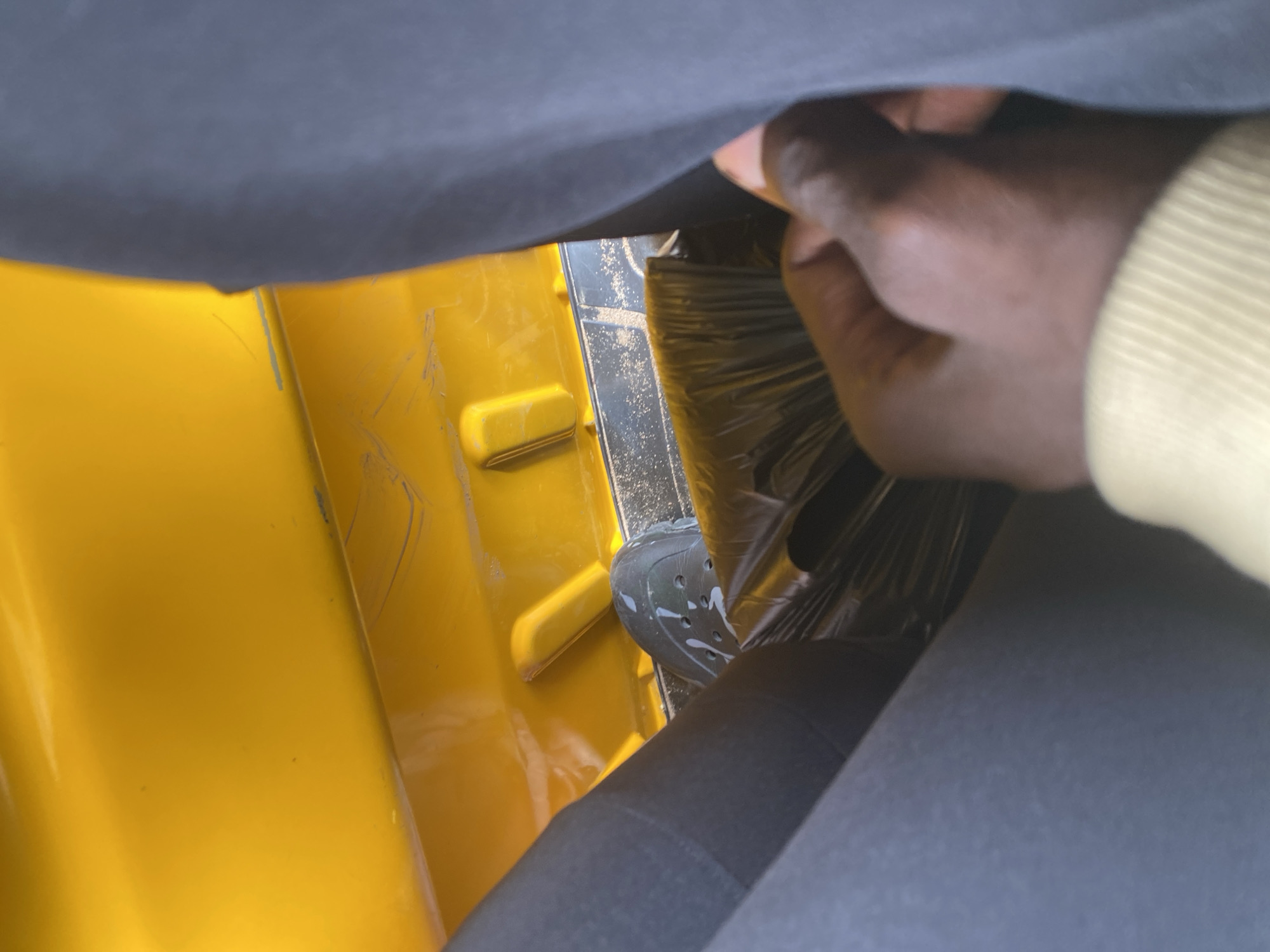This media file shows a close-up view of a person's hand reaching into a yellow interior, possibly a vehicle. The hand, with dark skin, is grasping a dark, plastic-wrapped object. Part of a black shoe or boot is visible on the floor, with white speckles on it. The setting appears to be inside a confined space, suggested by the limited view and the presence of upholstery or fabric in the foreground and background. The yellow color of the interior is prominent, indicating a brightly colored surface, possibly a painted metal panel. Some faint scratches or marks are visible on the yellow surface. The lighting appears to be natural, suggesting daylight. There is no visible text in the image. The context provided places this scene in Jalingo, Nigeria.