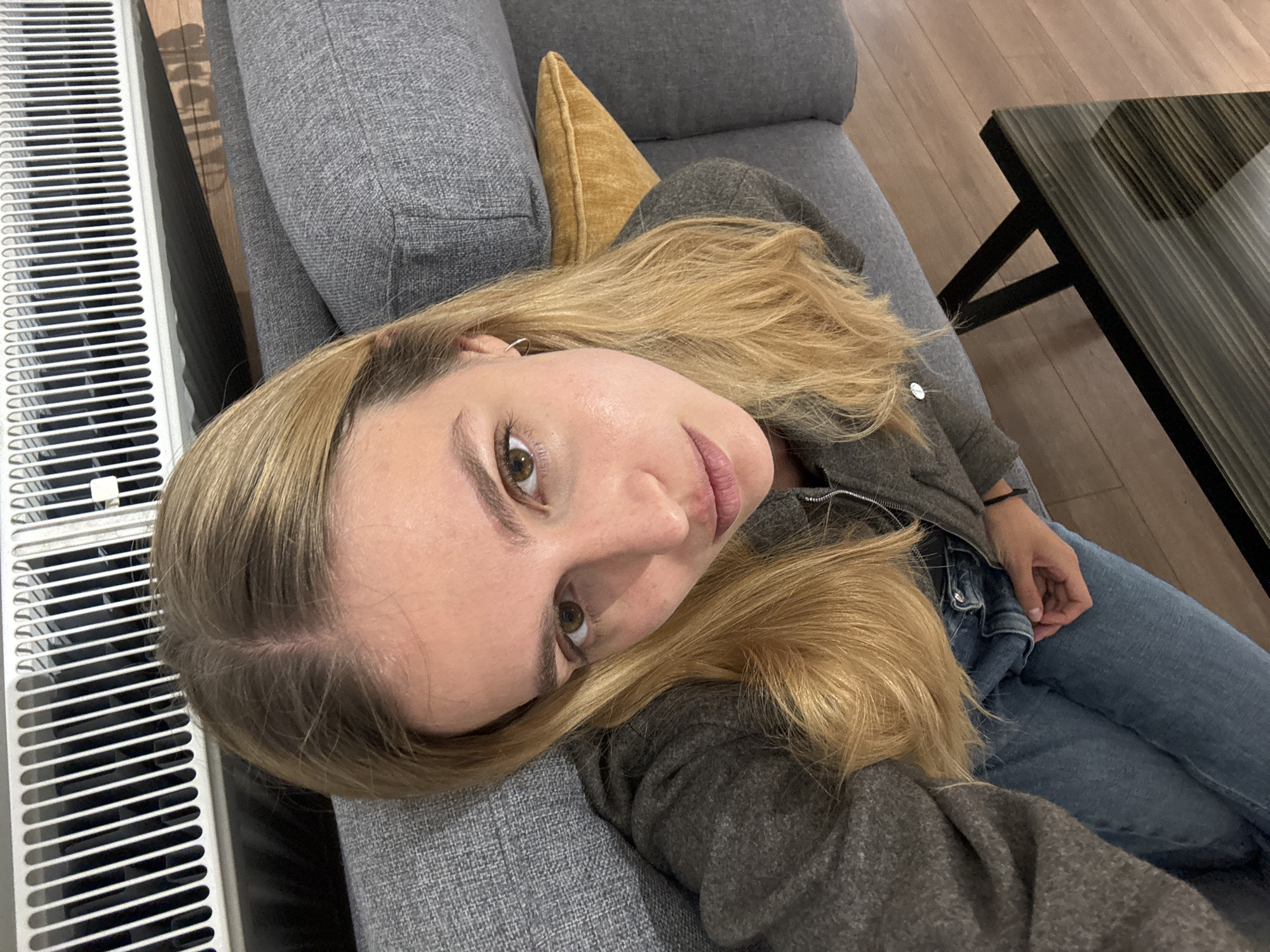A woman lies on a gray sofa with a mustard-colored pillow, wearing a gray jacket and blue jeans. Her blonde hair is styled loosely around her face. The environment includes a white radiator to the left and a dark, glass-topped coffee table in the background. The hardwood floor is visible. The photograph appears to be taken indoors. It is not possible to verify the location as Tbilisi, Georgia from the provided image.
