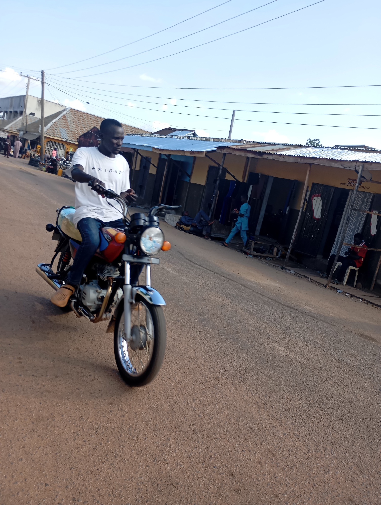 A man in a white t-shirt and blue jeans rides a motorcycle on a paved road in Shendam, Nigeria. The man is in the process of turning his head to his left as he drives, with his left hand on the handlebars. His t-shirt has "FRIENDS" written on it in black letters. The motorcycle is a red, blue, and chrome model with a visible engine and exhaust pipe. He is wearing brown sandals on his feet. Behind him, on the right side of the road, are a series of small shops with corrugated metal roofs. A person in a red shirt is sitting on a white plastic chair outside one of the shops, looking down at their phone. Another person in a blue outfit is walking away from the shops. In the background, there are utility poles with wires running across the sky, and buildings, including a water tower. The road is a reddish-brown asphalt, and the sky is a clear, light blue. The lighting suggests it is daytime, likely late afternoon due to the shadows.