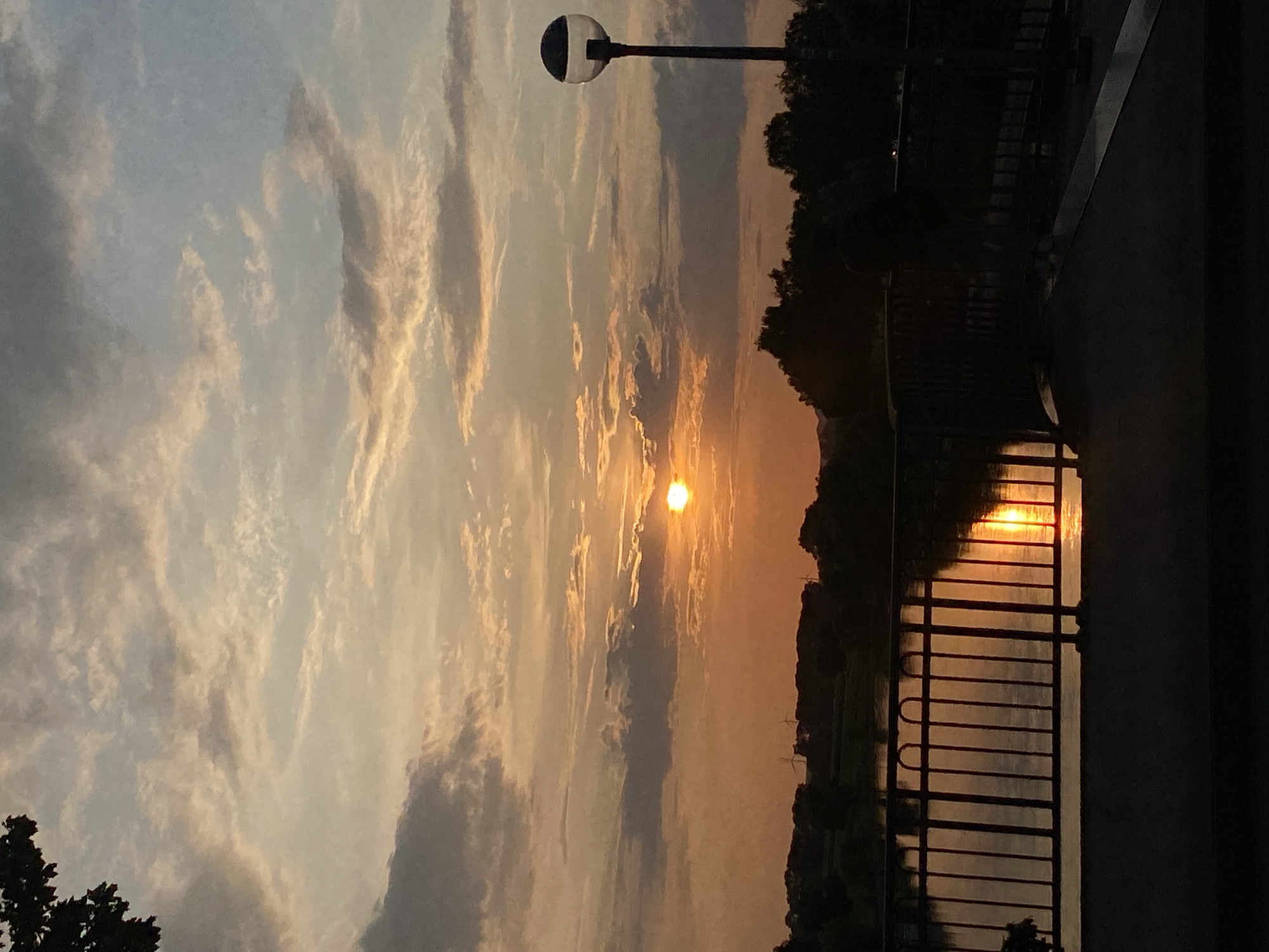 The image captures a sunset over the Weser River in Rinteln, Germany. The sky is filled with a blend of orange, yellow, and blue hues, with scattered clouds. The sun, a bright orb, is positioned just above the horizon, casting a warm glow across the scene. In the foreground, a dark silhouette of a bridge railing separates the viewer from the water. The river reflects the sunset's colors and the railing's structure. On the opposite bank, a line of trees creates a dark silhouette against the sky, with a few wind turbines visible in the distance. A modern lamppost with a spherical light fixture stands on the right side of the bridge. The overall mood is serene and tranquil, characteristic of a peaceful evening by the river.