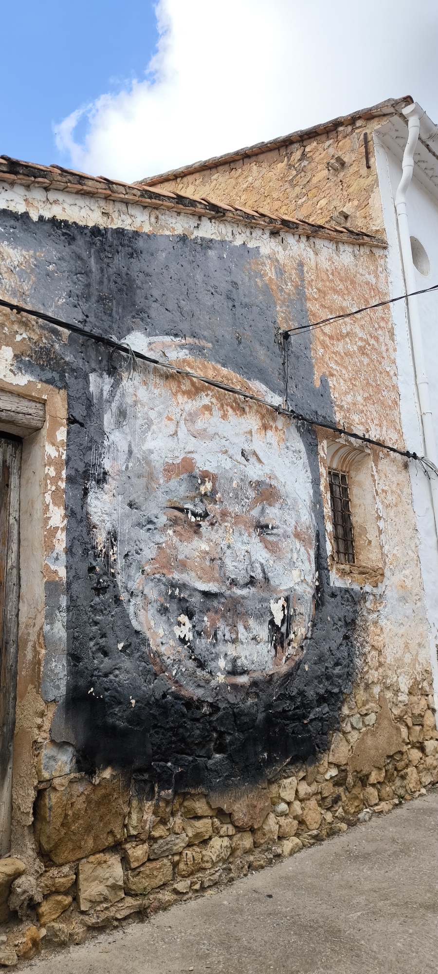 This is a photograph of a weathered wall in Fanzara, Spain, featuring a large, abstract graffiti portrait. The portrait, rendered in shades of black, white, and brown, appears to depict a face with closed eyes and a wide smile. The paint is peeling and distressed, revealing the rough texture of the stone and plaster wall beneath. The wall itself shows signs of age and decay, with chipped paint and crumbling sections. A small, barred window is visible to the right of the graffiti, and electrical wires run horizontally across the wall. The lower portion of the wall is constructed from rough-hewn stones. The ground in front of the wall is a paved surface, likely a street or sidewalk, and the background shows a portion of a building with a tiled roof and a white downspout under a blue sky with white clouds.