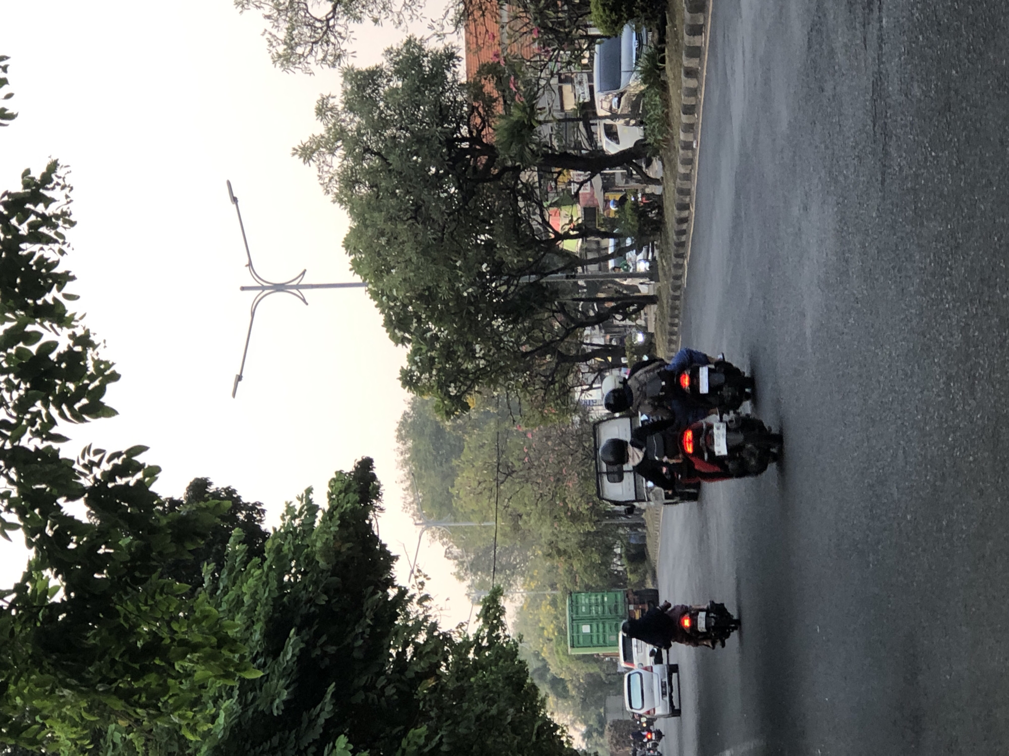 The image shows a street scene in Sumorame, Indonesia. Multiple people are riding motorcycles on a paved road, with some wearing helmets and carrying backpacks. Various vehicles, including cars and a green shipping container on a truck, are also present. The road is lined with trees and there are buildings visible in the background. The sky is overcast, suggesting it could be early morning or late afternoon. The overall impression is that of a typical urban or suburban environment in Indonesia.