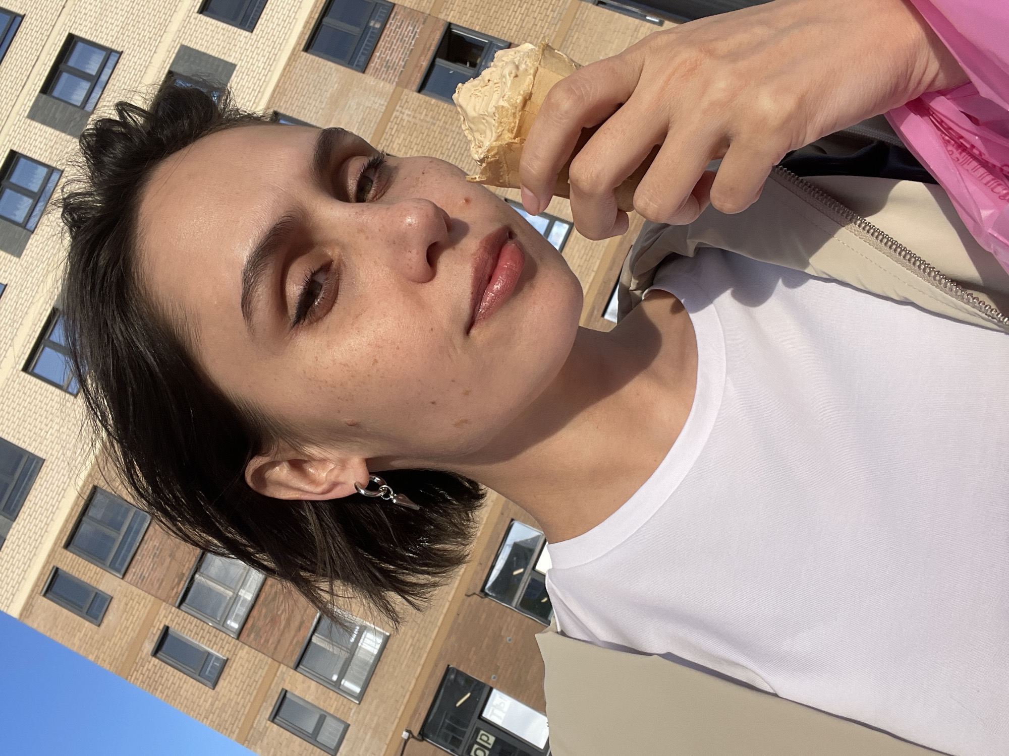 A young woman with short dark hair and freckles is holding an ice cream cone in front of her face. She is wearing a white t-shirt and a light-colored jacket. The background shows a tall brick building with numerous windows. The sky is visible in the lower-left corner and appears to be clear and blue. The location is Izhevsk, Russia.