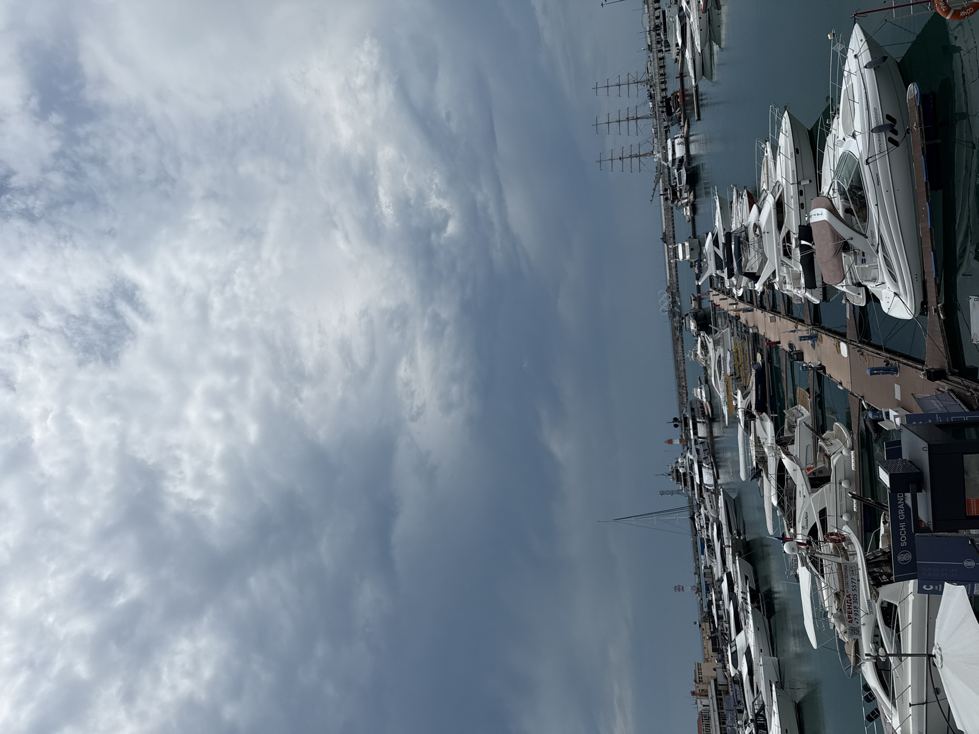 The image depicts a marina filled with numerous yachts and boats, situated in the city of Sochi, Russia. The sky above is overcast with dark, dramatic clouds, suggesting an impending storm or a very moody day. The water in the marina is calm and reflects the sky and the vessels. The yachts are of various sizes, with many being sleek, modern vessels. In the background, larger sailing ships with multiple masts are visible, along with some port infrastructure and buildings. A prominent sign reads "SOCHI GRAND," indicating the location. There are no visible people actively engaged in activities within the frame of the shot. The overall scene is one of a well-equipped marina under a heavy, cloudy sky.