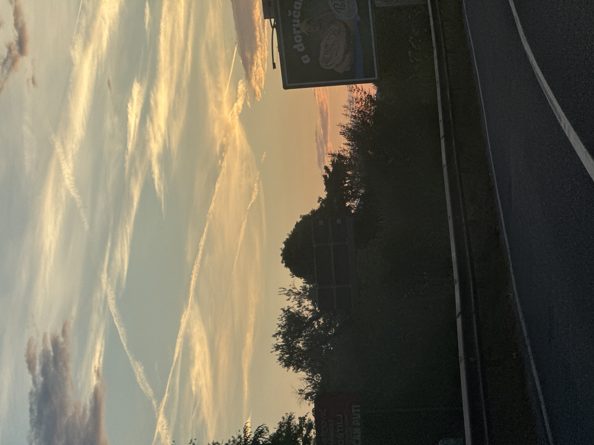 The image depicts a sunset over a roadside in Kamenovo, Montenegro. The sky is filled with wispy clouds and contrails, illuminated by the warm hues of the setting sun, ranging from soft orange to yellow. In the foreground, a dark asphalt road with white lane markings stretches into the distance. To the left of the road, a dense line of trees and foliage forms a dark silhouette against the vibrant sky.
A billboard is visible on the right side of the image, partially obscured by trees. The text on the billboard is mostly illegible due to the low light and angle, but some words are discernible, appearing to be in a Slavic language. One part of the text reads "a doručak" and another partially visible section appears to be "PUTI". The overall mood is serene, with the natural beauty of the sunset contrasting with the man-made elements of the road and billboard. The time of day is clearly evening, as indicated by the sunset colors and the diminishing light. The weather appears to be clear, with the clouds contributing to the picturesque sky.