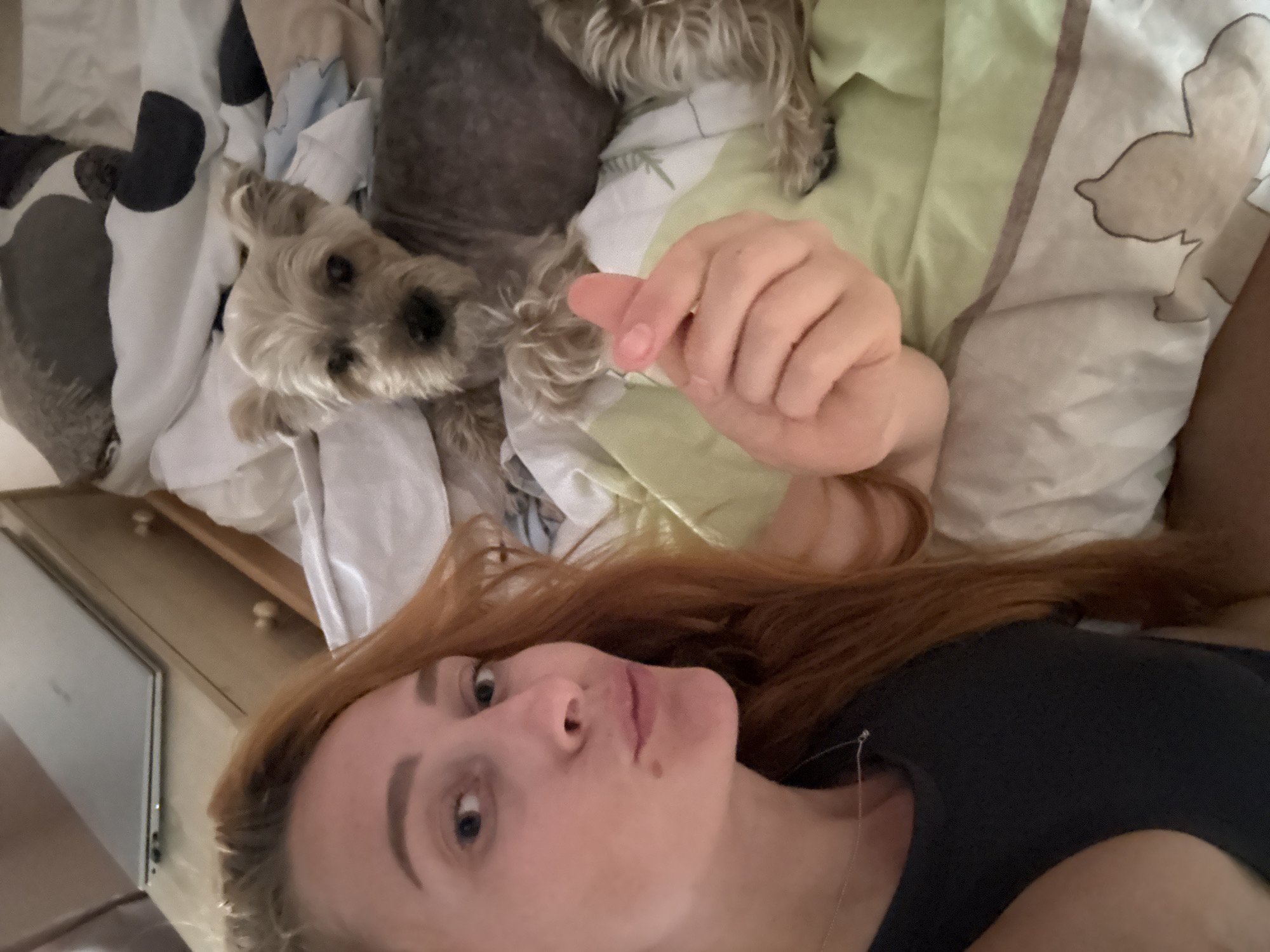 In Budva, Montenegro, a woman with long, reddish-brown hair is captured in a selfie lying in bed. She is wearing a dark, sleeveless top and a delicate silver necklace. Her left hand is raised, forming a heart shape with her thumb and index finger. Beside her, on the bed, a small, light-brown terrier-type dog is lying down. The bed is made with a white duvet featuring a light green and brown floral pattern. In the background, a wooden dresser with two drawers is visible, topped with a closed silver laptop. The overall scene depicts a relaxed, intimate moment between the woman and her pet.