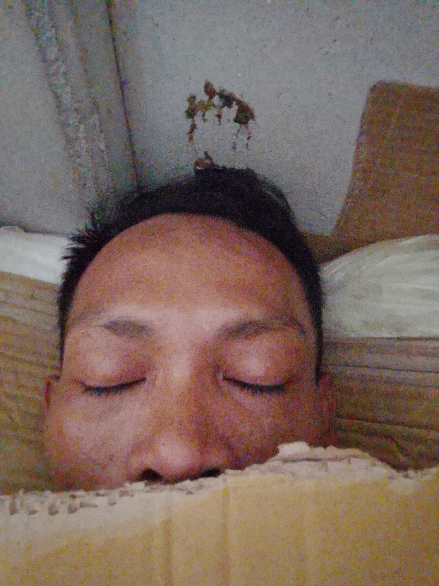 A close-up shot shows a person with closed eyes lying down. Only their face and the top of their head are visible. The person has dark hair and a reddish skin tone. They are partially covered by a yellow blanket or fabric. The background includes a white pillow and a ceiling with discoloration and a spot of what appears to be organic material. The location is Bukit Ketok, Indonesia.