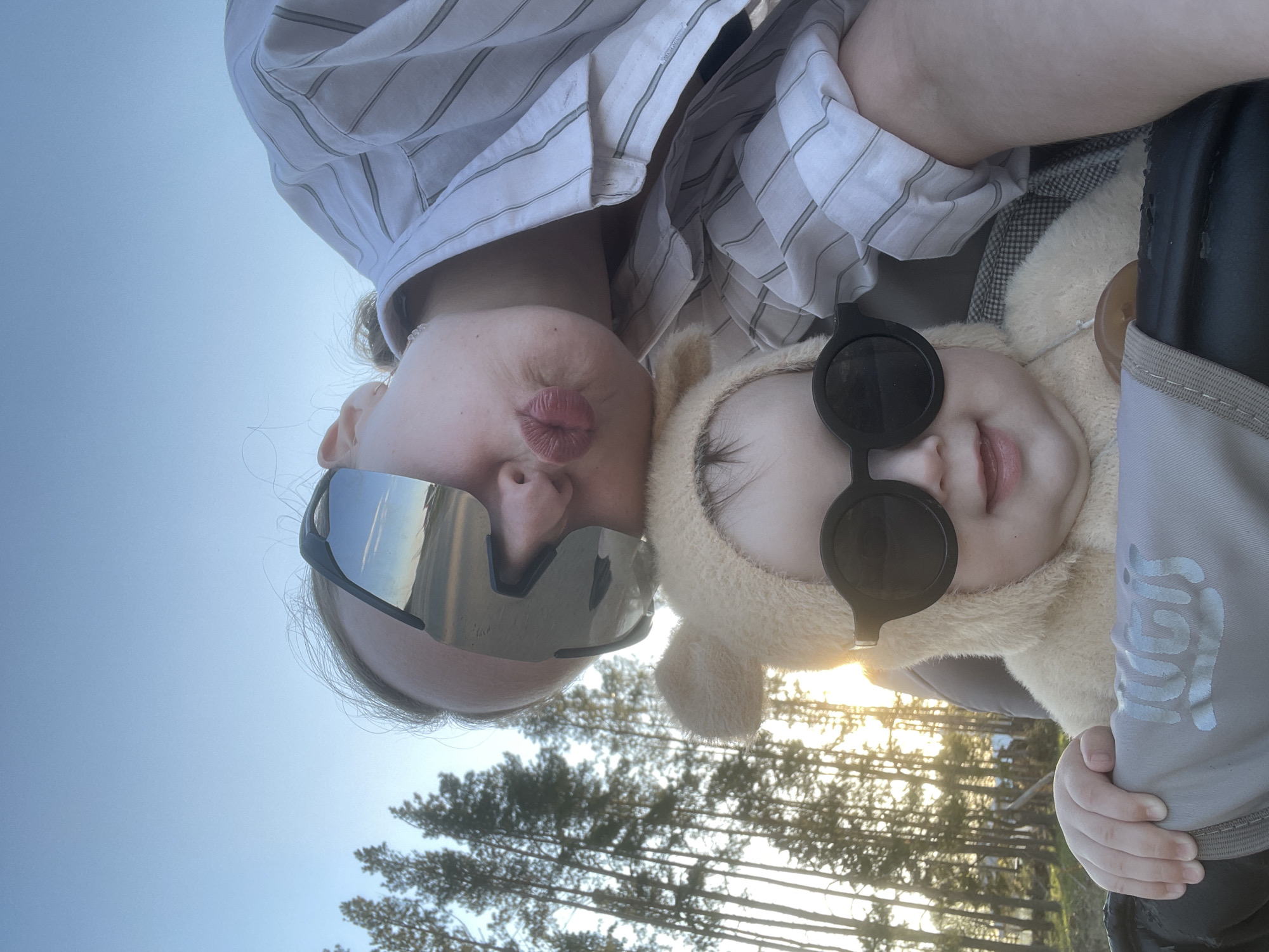 The image depicts a selfie taken in Podshivalovo, Russia. A person, presumably an adult, is wearing reflective sunglasses and a striped shirt. They are pursing their lips, possibly in a kissing gesture. Next to them is a baby wearing a furry hooded outfit and sunglasses. The baby is being held in a carrier. The background includes trees and a clear blue sky with sunlight filtering through the trees.
